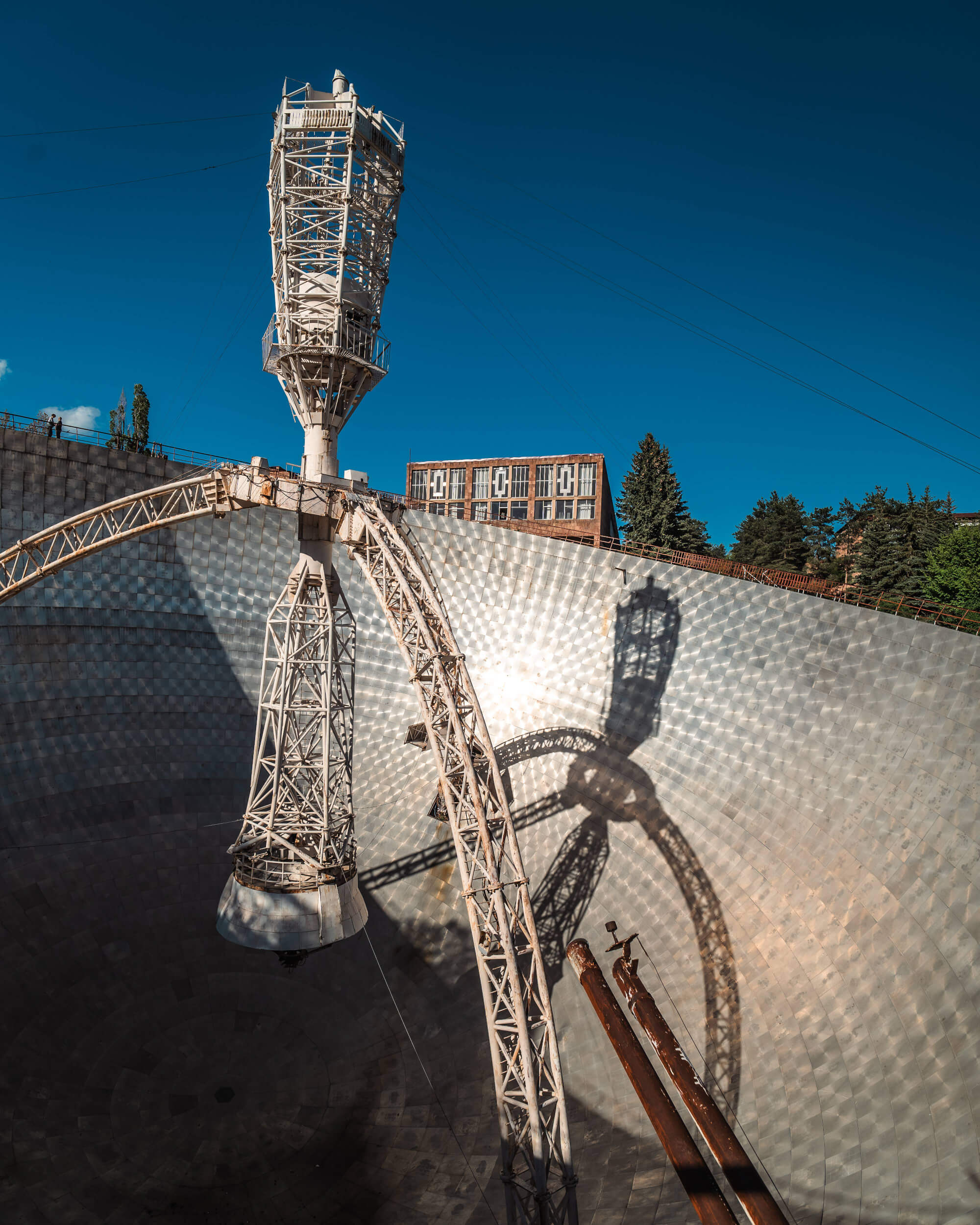 An industrial structure with a tall tower and a curved metallic base, casting a complex shadow on a textured surface. The background features trees and a building against a bright blue sky.