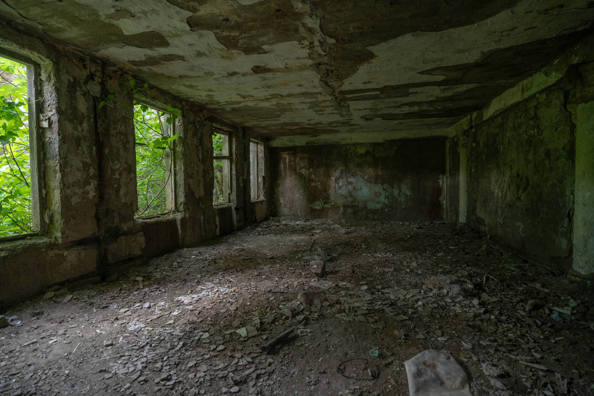 An abandoned room with peeling walls, scattered debris on the floor, and green foliage visible through the windows.