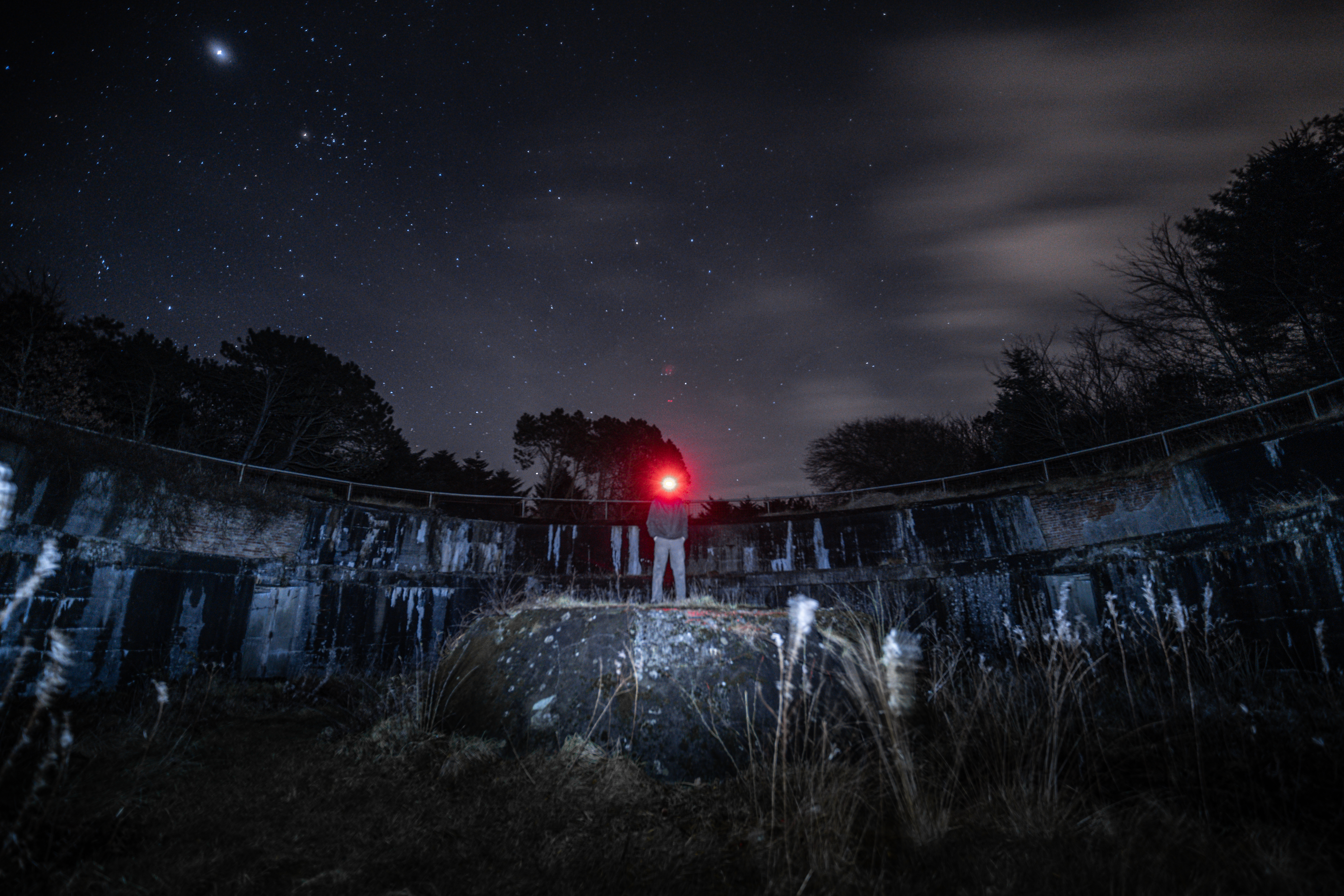 A person with a red flashlight stands on a stone inside a dark circular structure, surrounded by grass and trees, under a starry night sky.