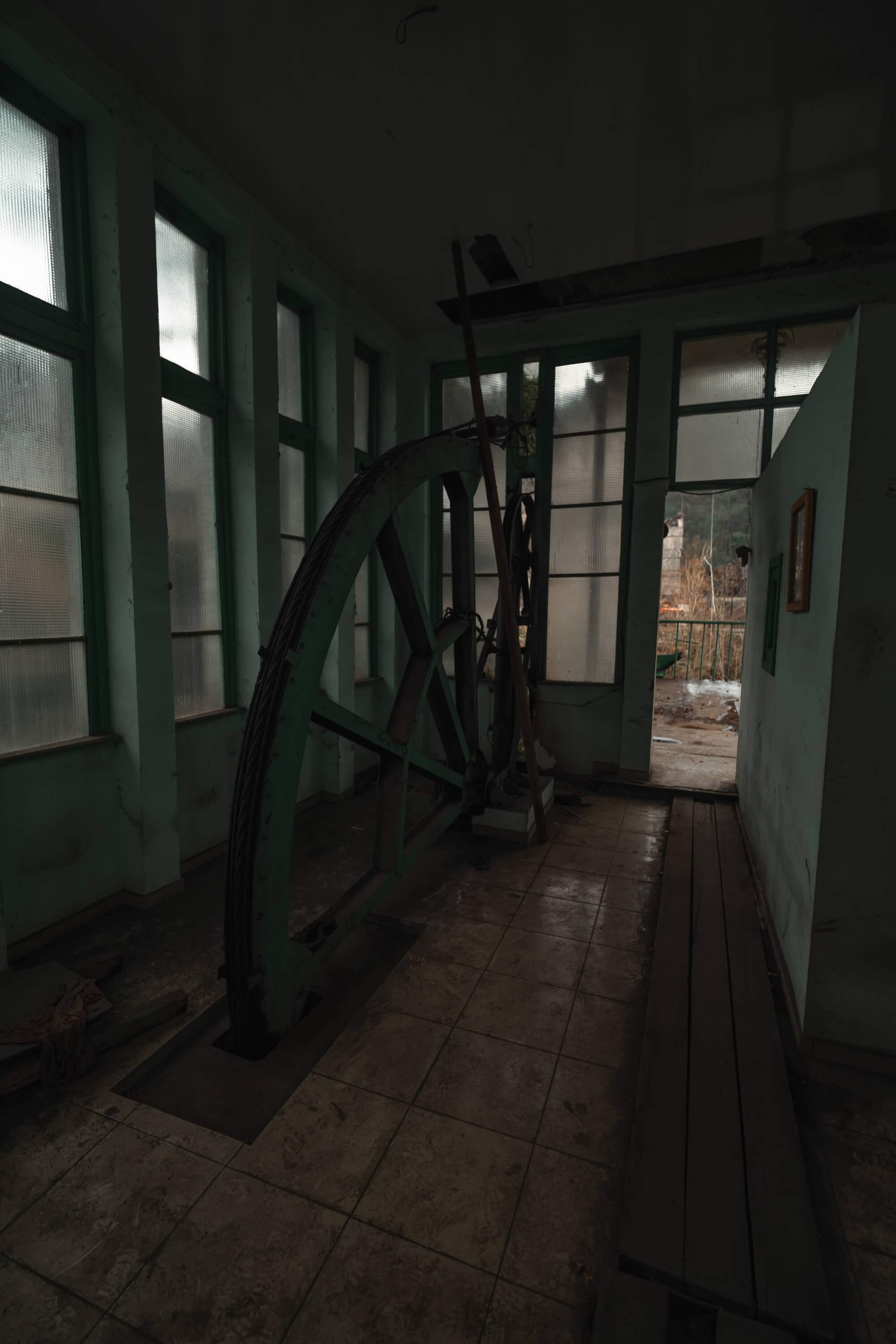 Interior of an abandoned industrial building showing a large green wheel, dusty windows, and a doorway leading outside.