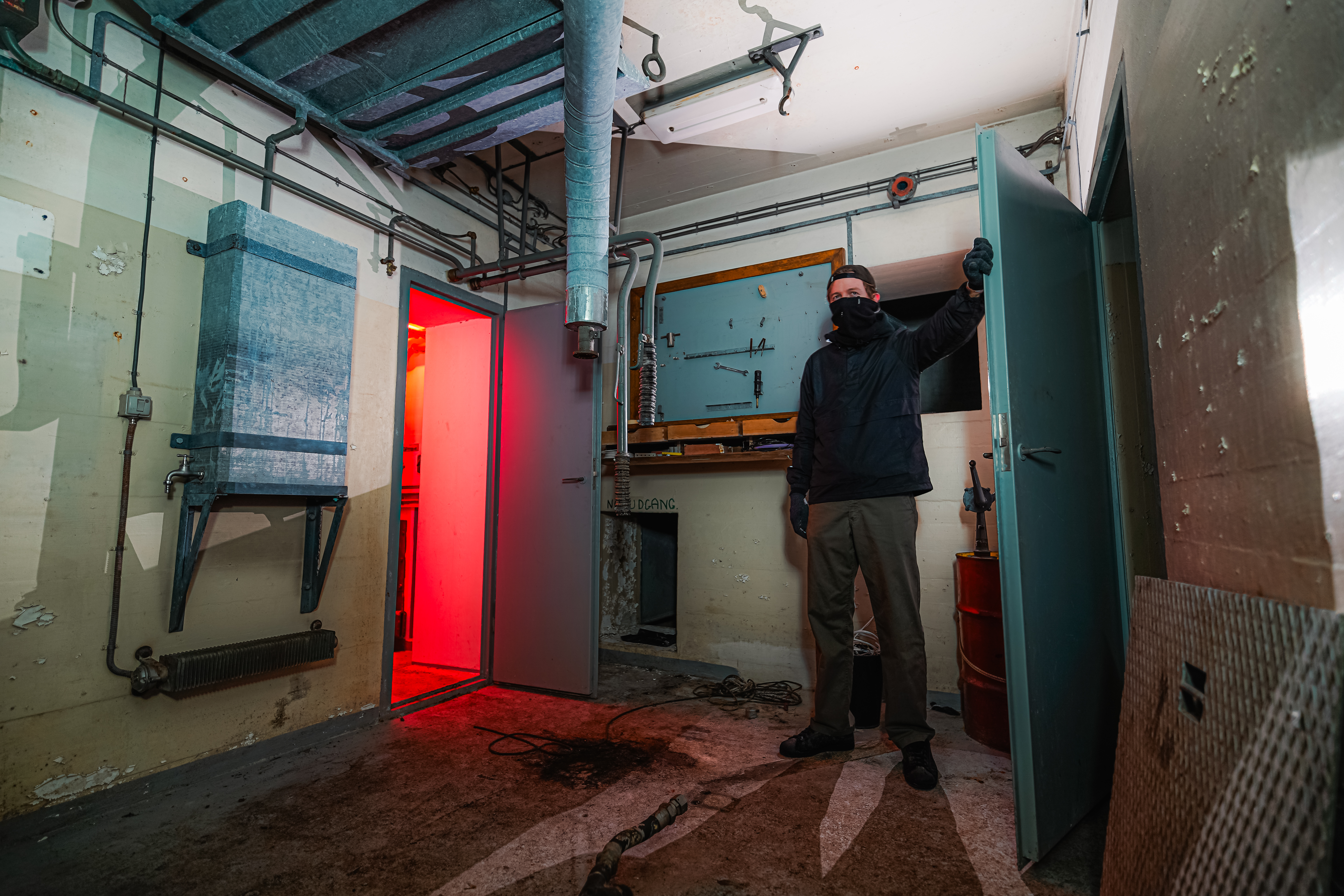 A masked person in a dark jacket stands near an open door emitting a red light in a dim, industrial room filled with pipes and scattered debris.