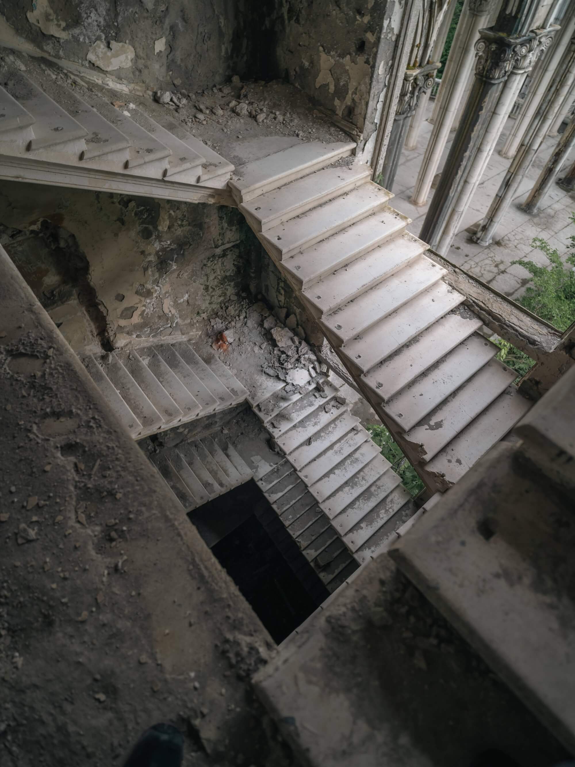 A top-down view of a decaying staircase with dusty white steps and dark shadows below, surrounded by cracked walls and remnants of an old structure.