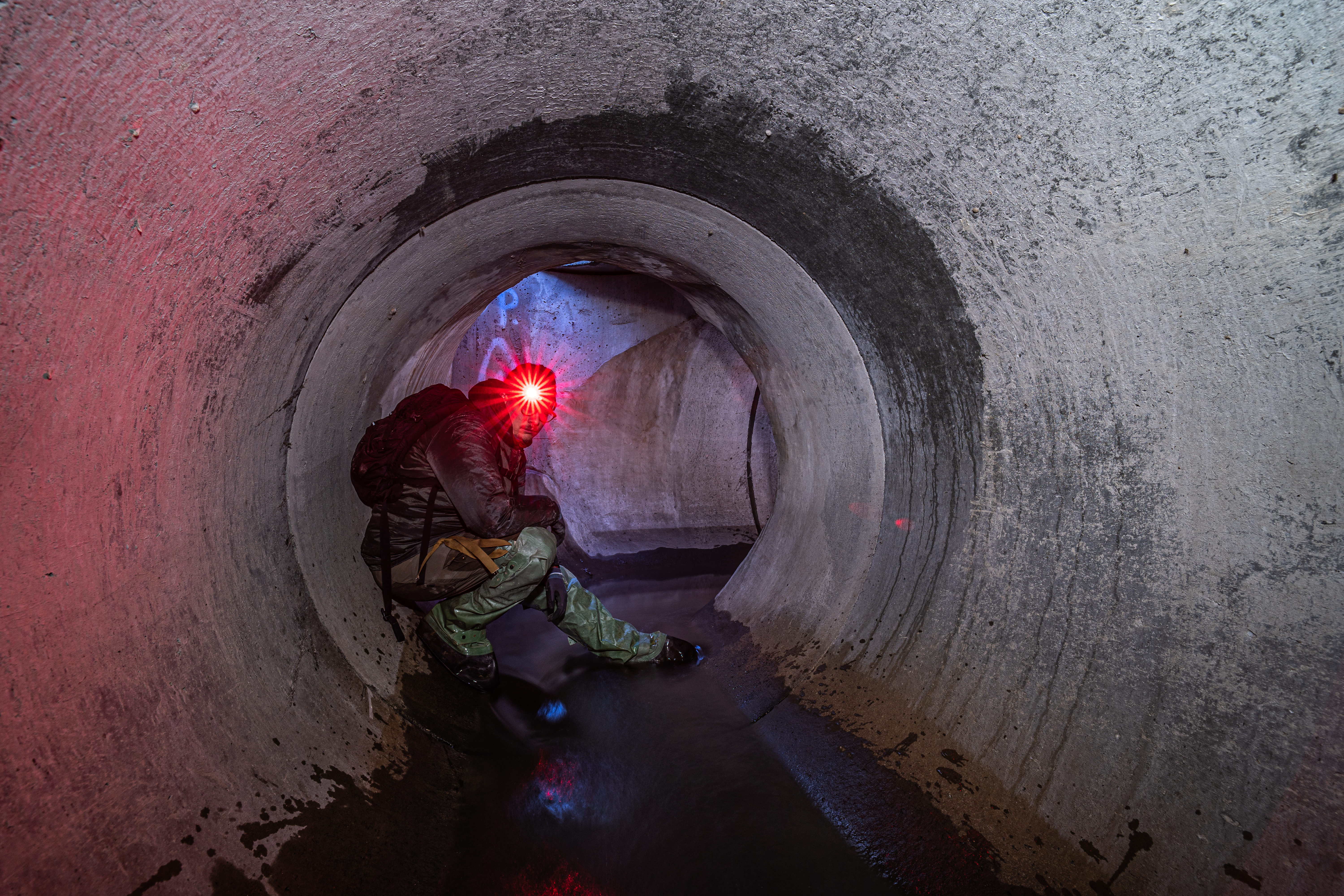 A person with a red headlamp sitting in a large, circular concrete tunnel filled with water, surrounded by textured walls reflecting various colors.