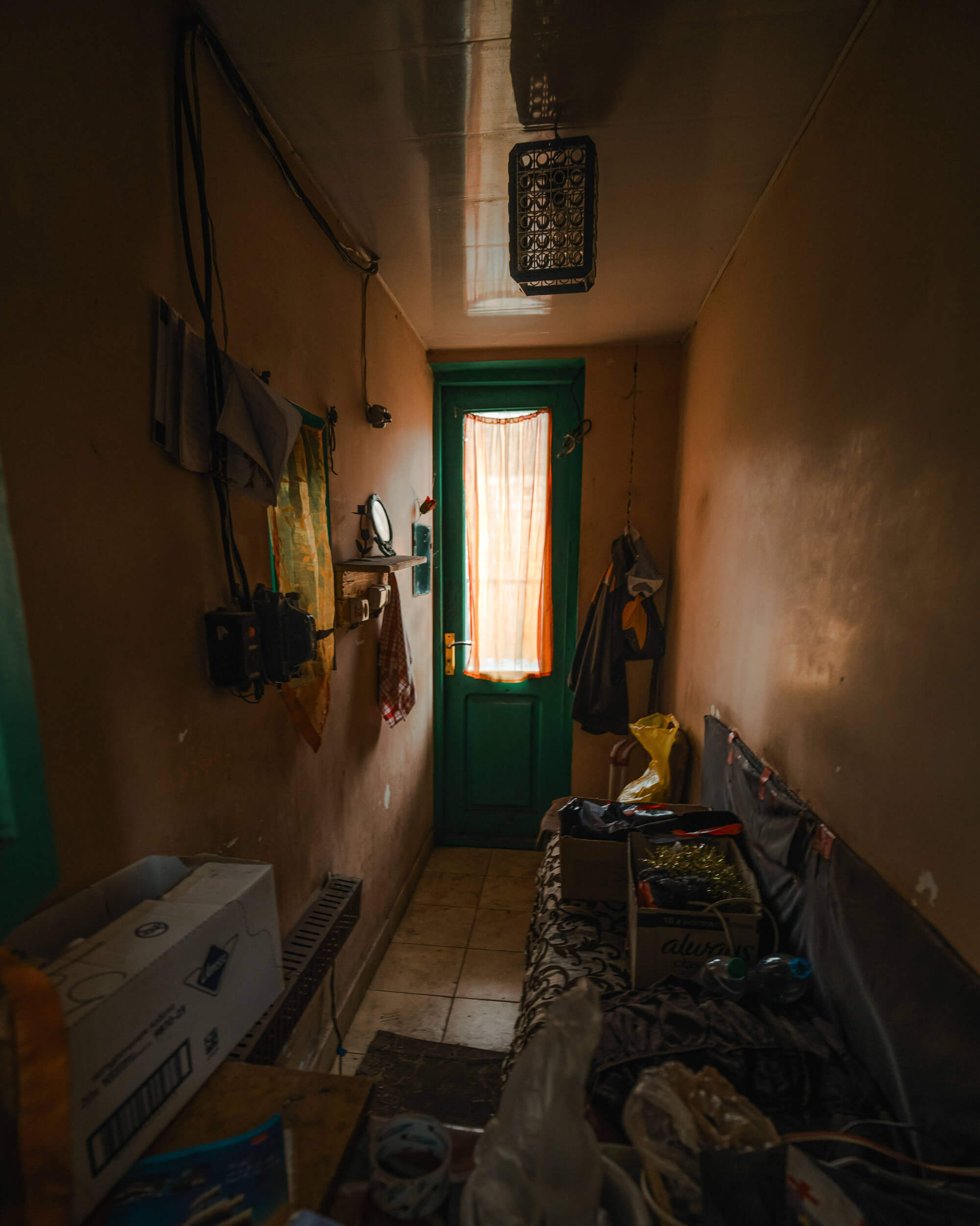A narrow hallway with beige walls and a green door at the end. Light filters through a sheer curtain over the door. A box and various items are scattered on a surface along the walls.