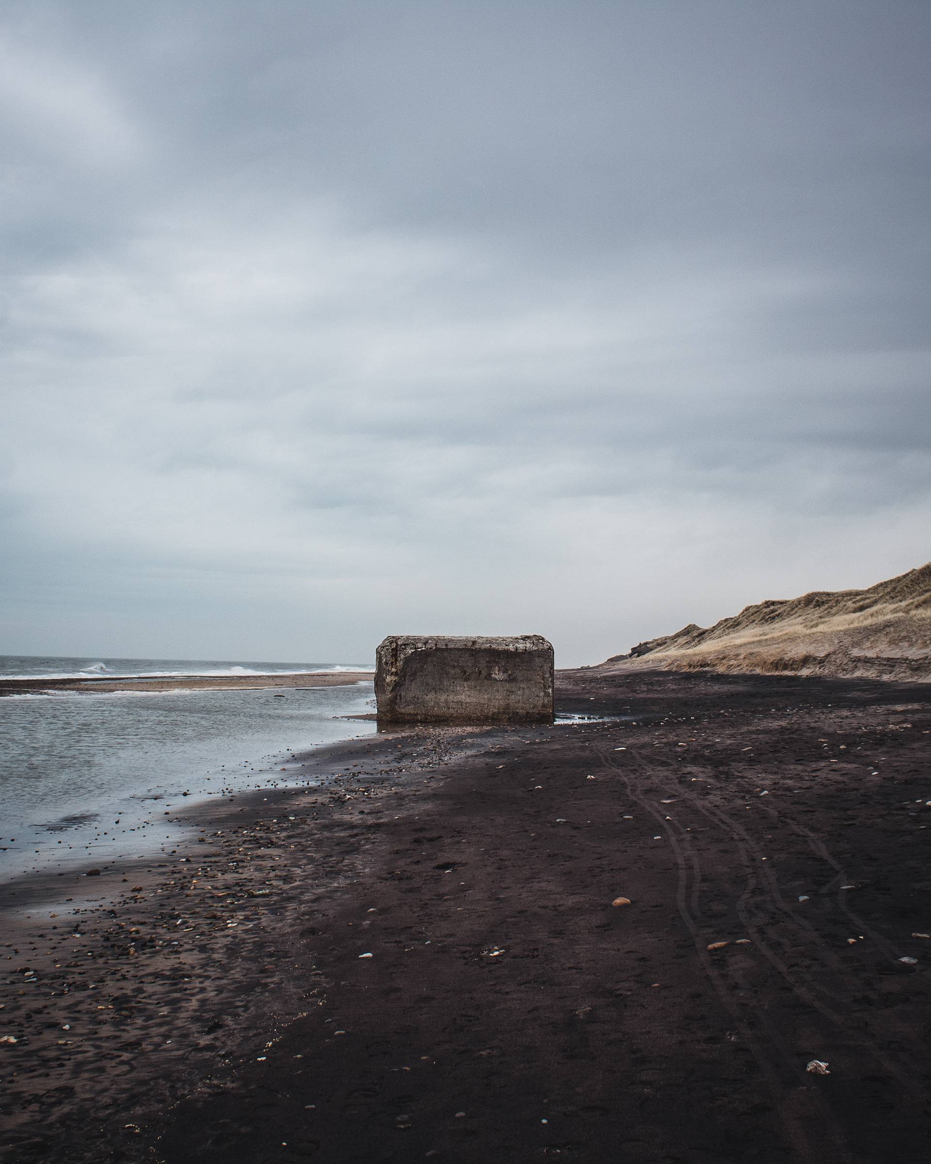A large, weathered concrete structure on a dark sandy beach with a gray, overcast sky above. Gentle waves are visible along the shore.
