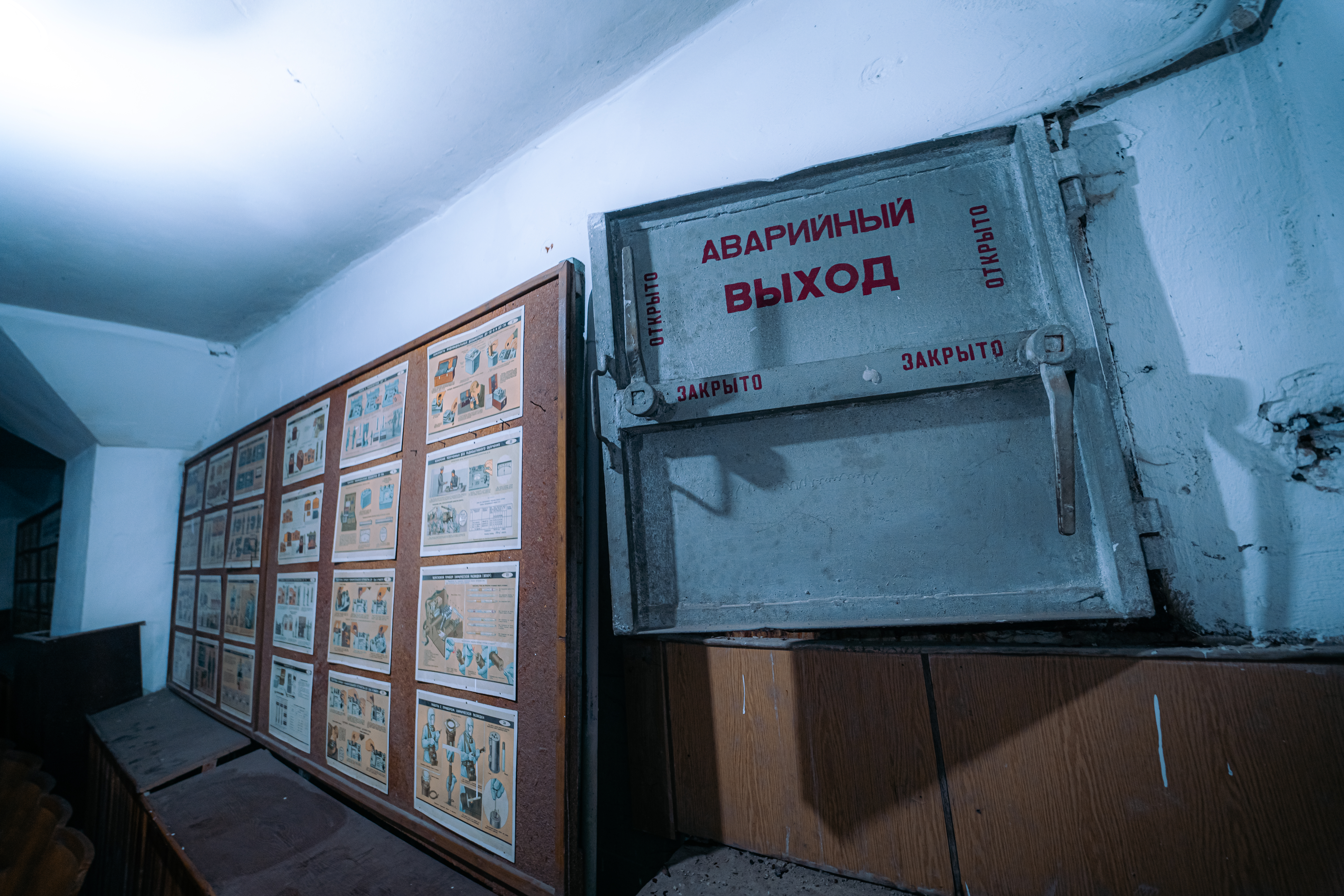 A dimly lit corridor with an emergency exit door on the right side and safety instruction posters on the left wall.