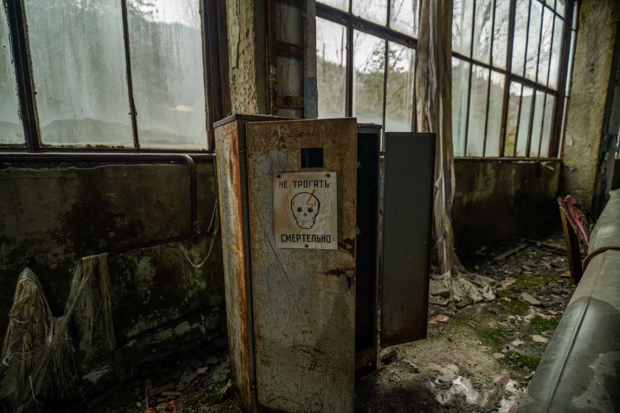 A rusty metal cabinet with a skull warning sign in an abandoned industrial space, featuring large grimy windows and scattered debris on the floor.