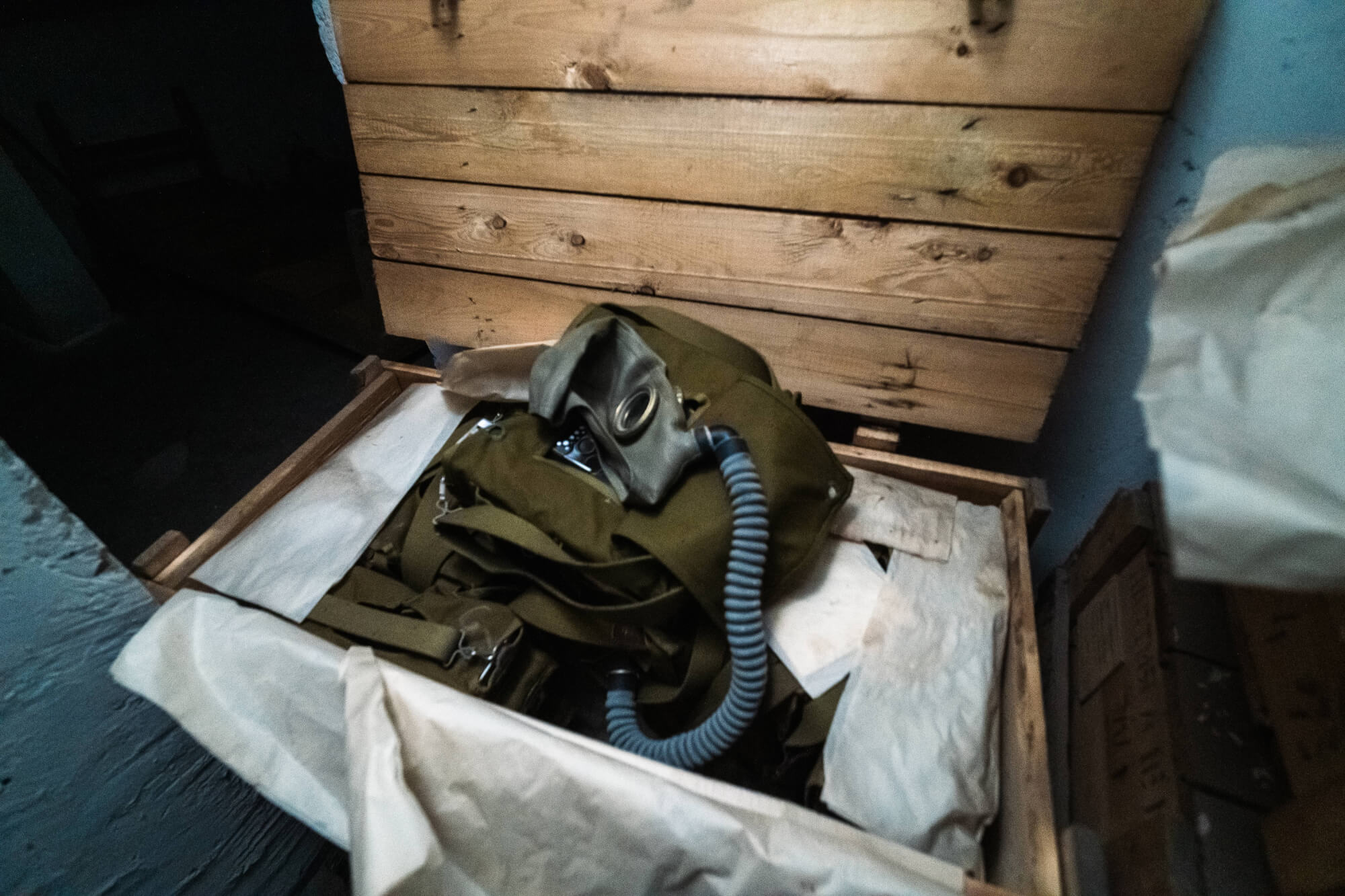 A wooden crate partially open revealing a green military backpack with a breathing apparatus inside, surrounded by crumpled white paper, set against a dark background.