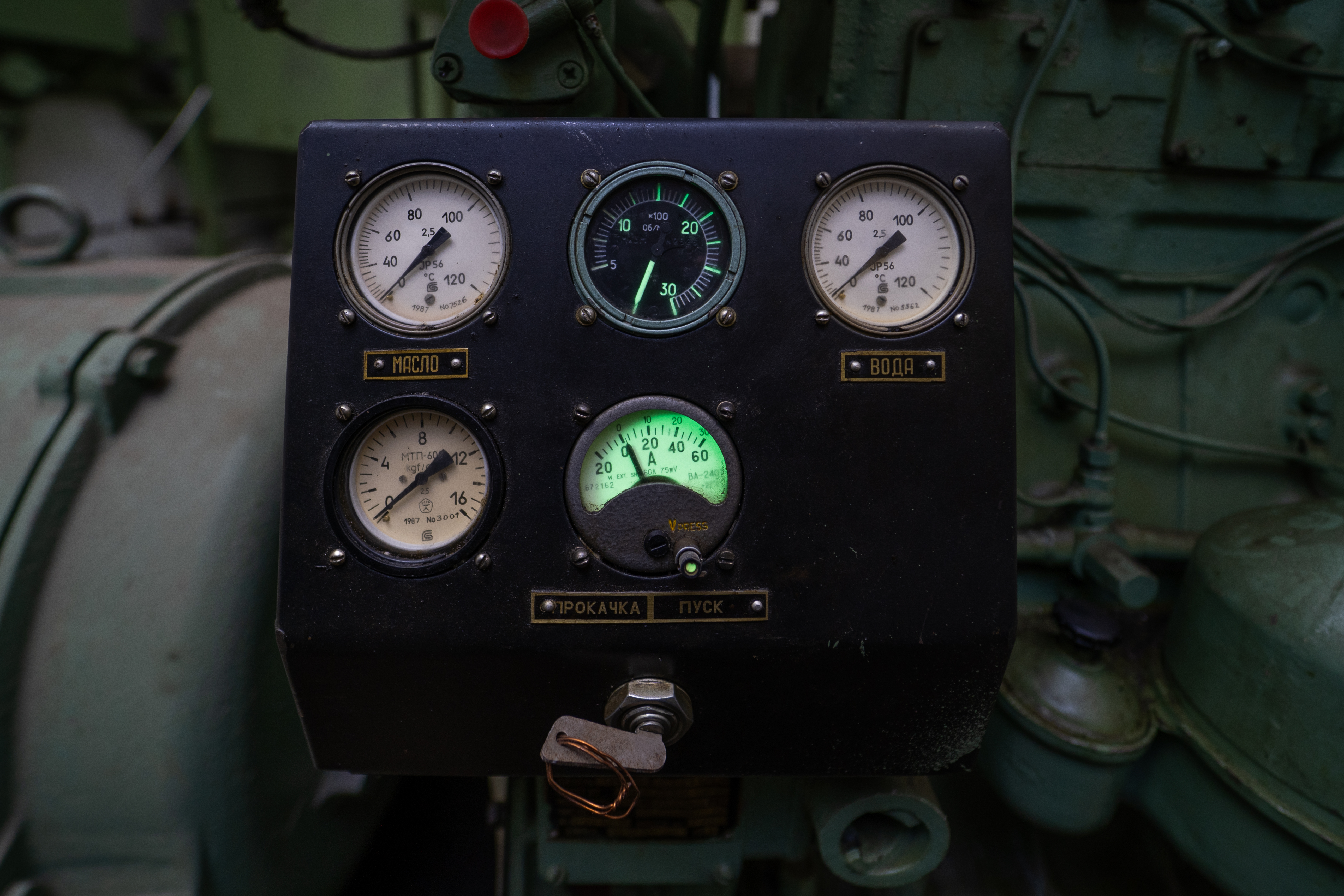 Close-up view of a vintage industrial control panel featuring several analog gauges for oil and water pressure, with one illuminated gauge showing a green light. The background includes bits of machinery, emphasizing a mechanical environment.