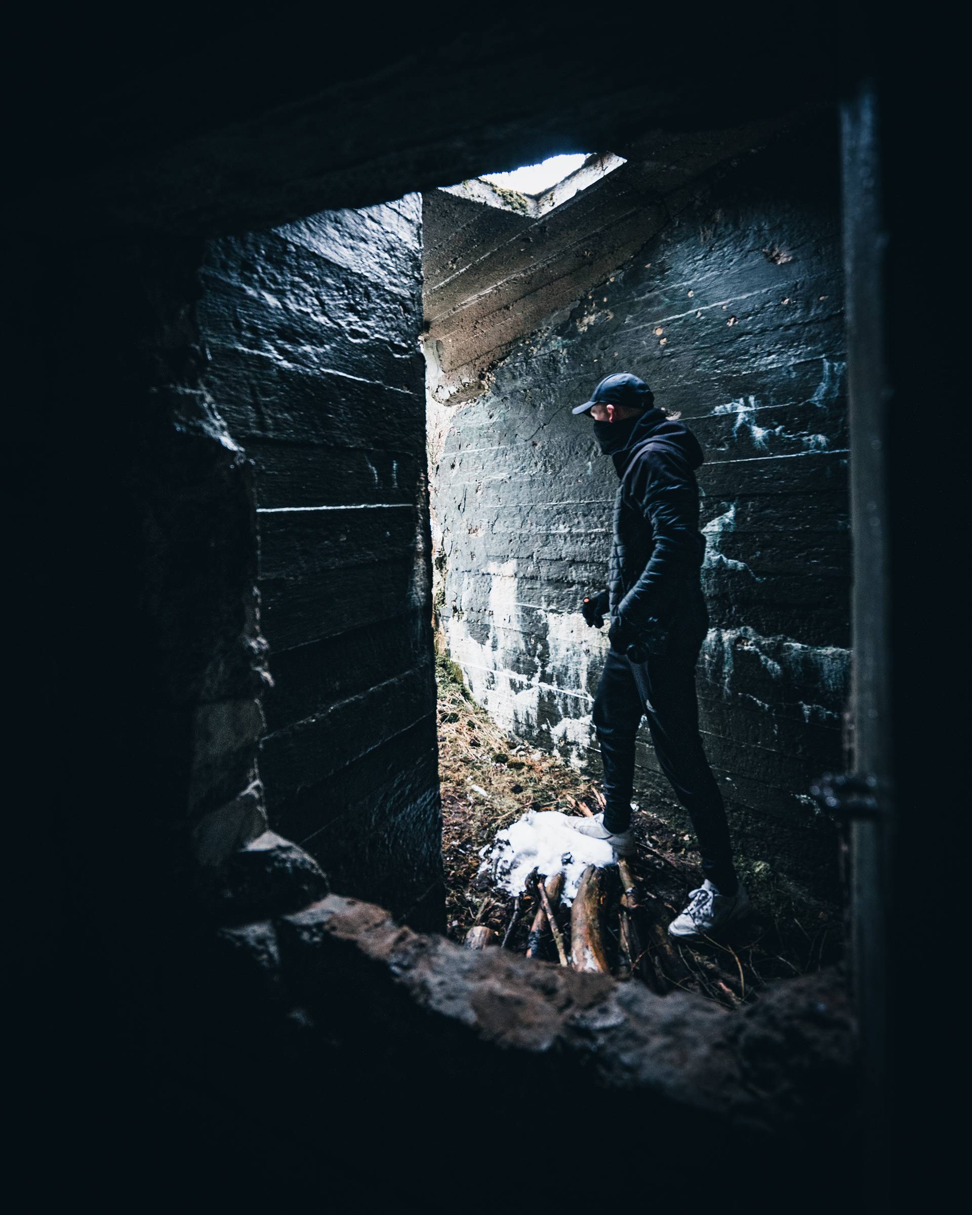 A person in dark clothing stands on a pile of logs covered in snow, viewed through an opening in a stone wall. The background consists of rough, dark stone walls and a glimpse of light from above.