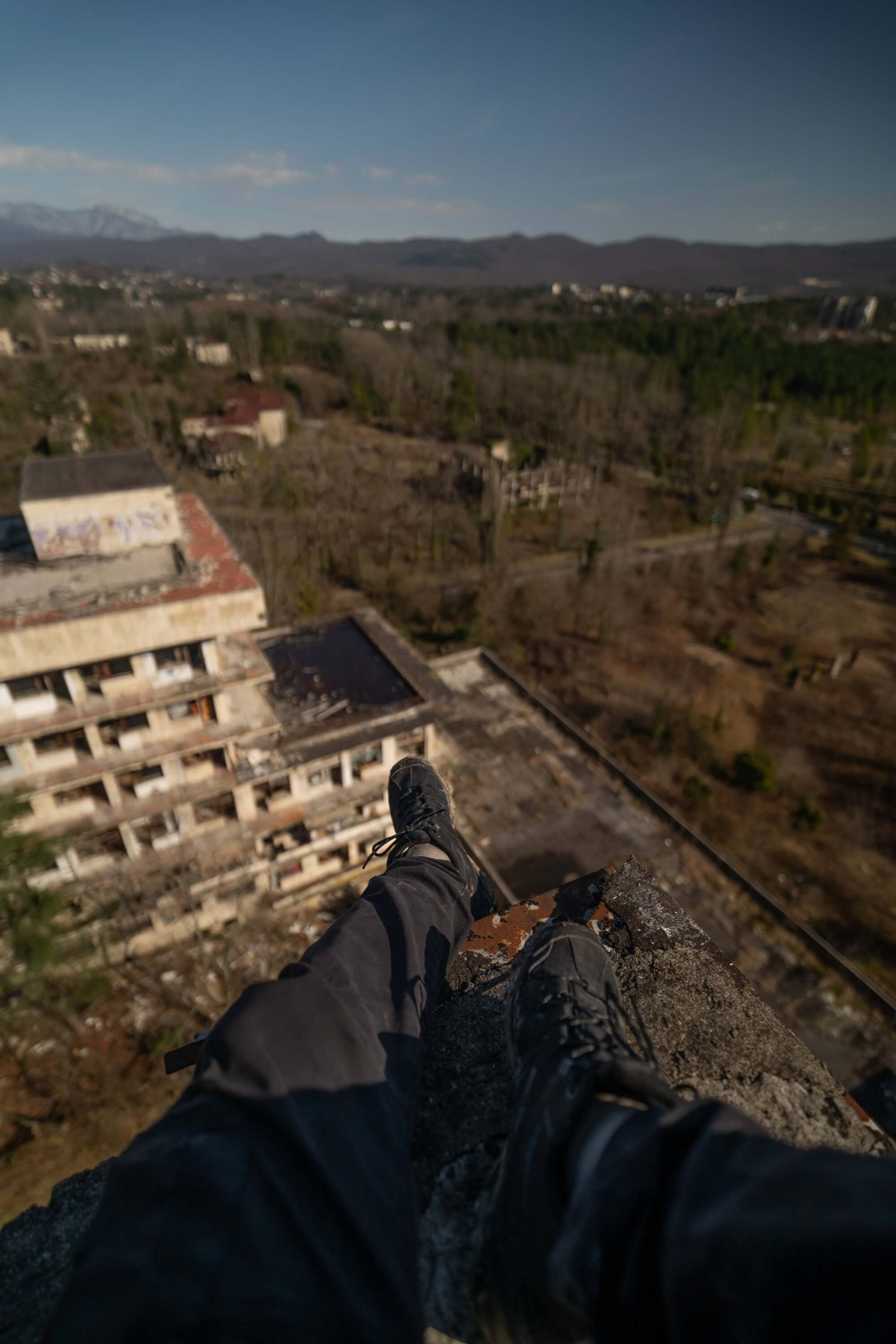 A pair of black boots and pants are positioned at the edge of a crumbling rooftop, looking down at an overgrown urban landscape with abandoned buildings and distant mountains under a clear blue sky.