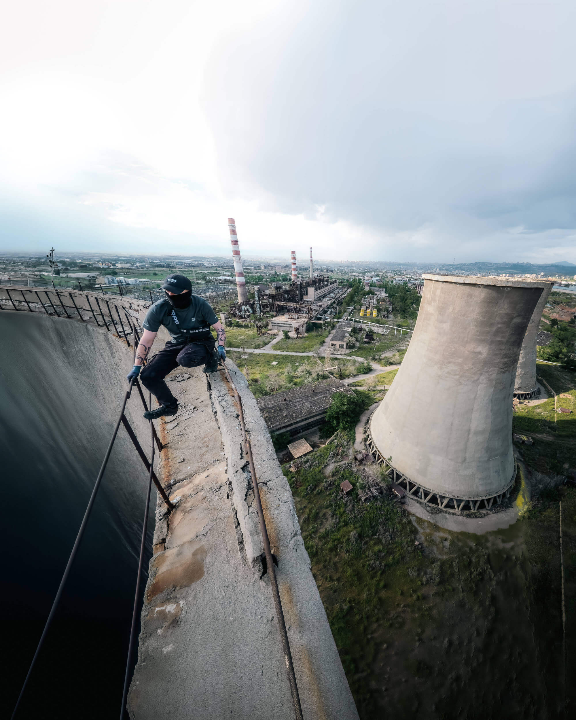 A person in a dark outfit and face covering stands on the edge of a crumbling concrete structure, with industrial cooling towers and abandoned factories visible in the background under a cloudy sky.