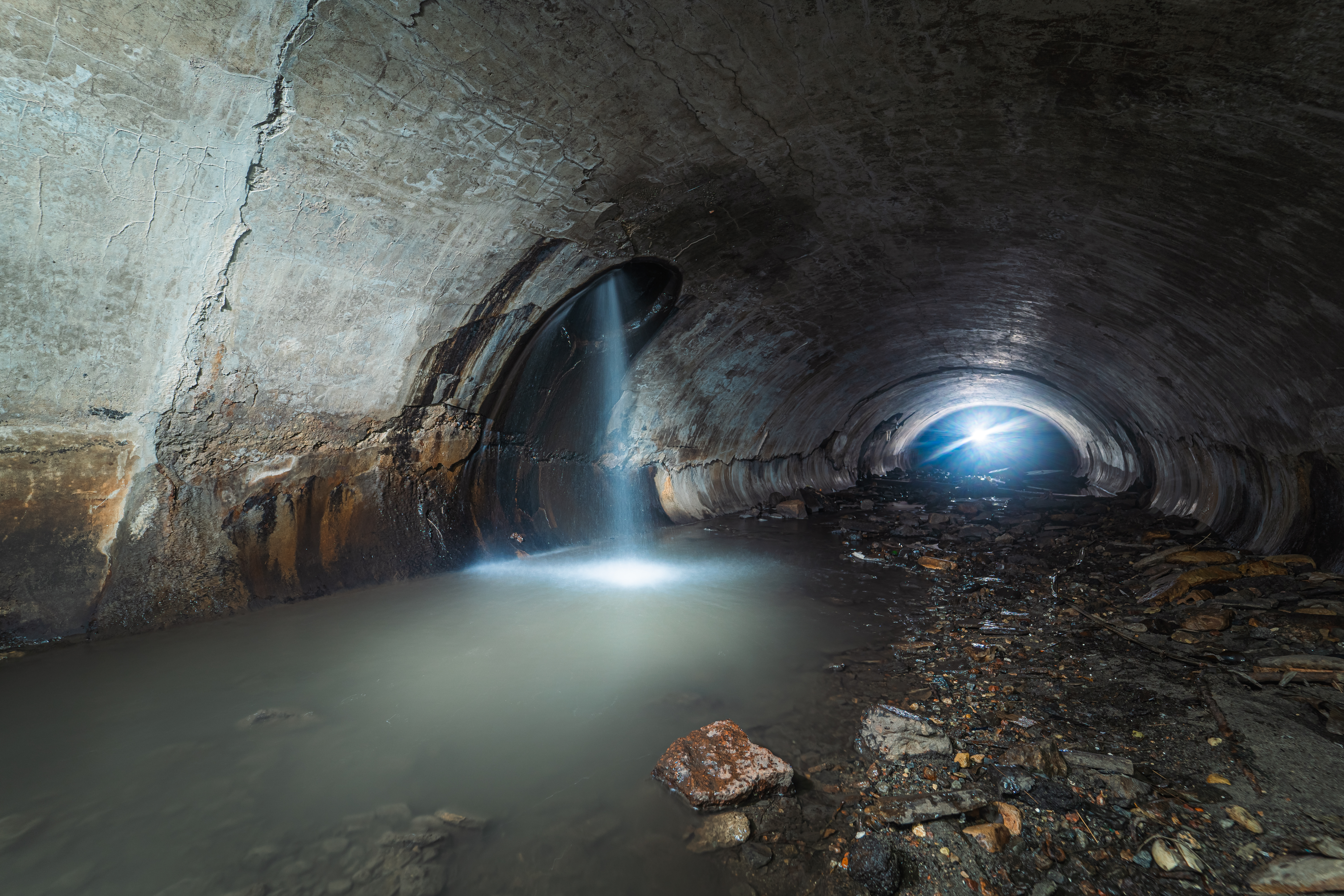 A spacious concrete tunnel with cracked walls, featuring a stream of water flowing from one opening. The floor is covered with stones and debris, while a distant bright light is visible at the opposite end of the tunnel.