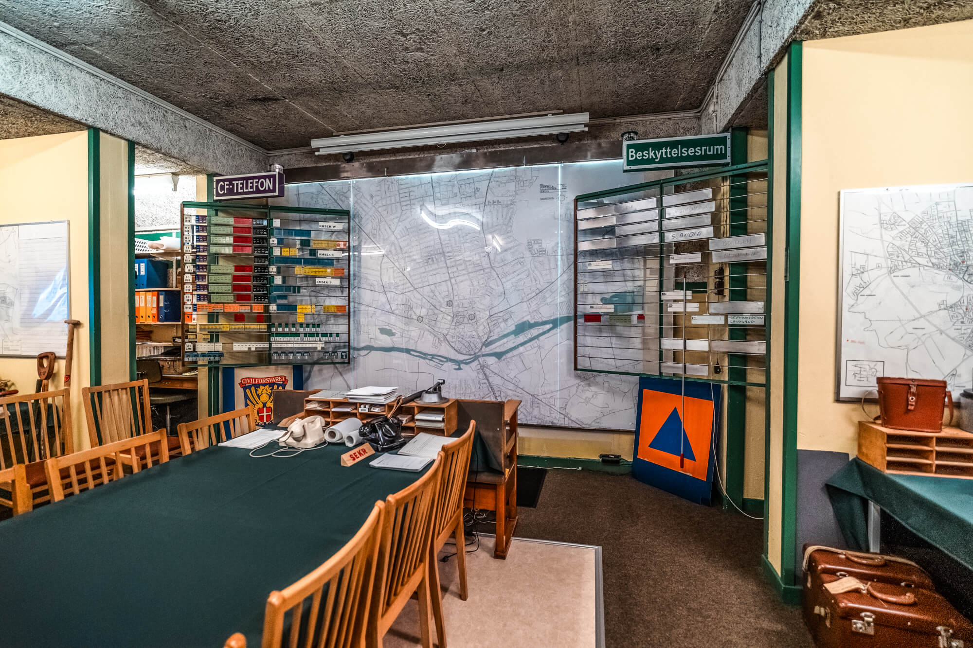 A vintage command center interior featuring a long table with wooden chairs, a wall-mounted map, organized index cards, and retro communication devices, all set against a textured concrete ceiling.