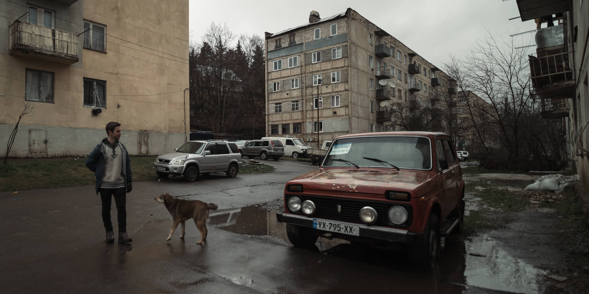 A young man stands beside a vintage red car in a rainy urban setting, where a dog walks nearby. In the background, worn-out apartment buildings and a gray sky set a somber mood.