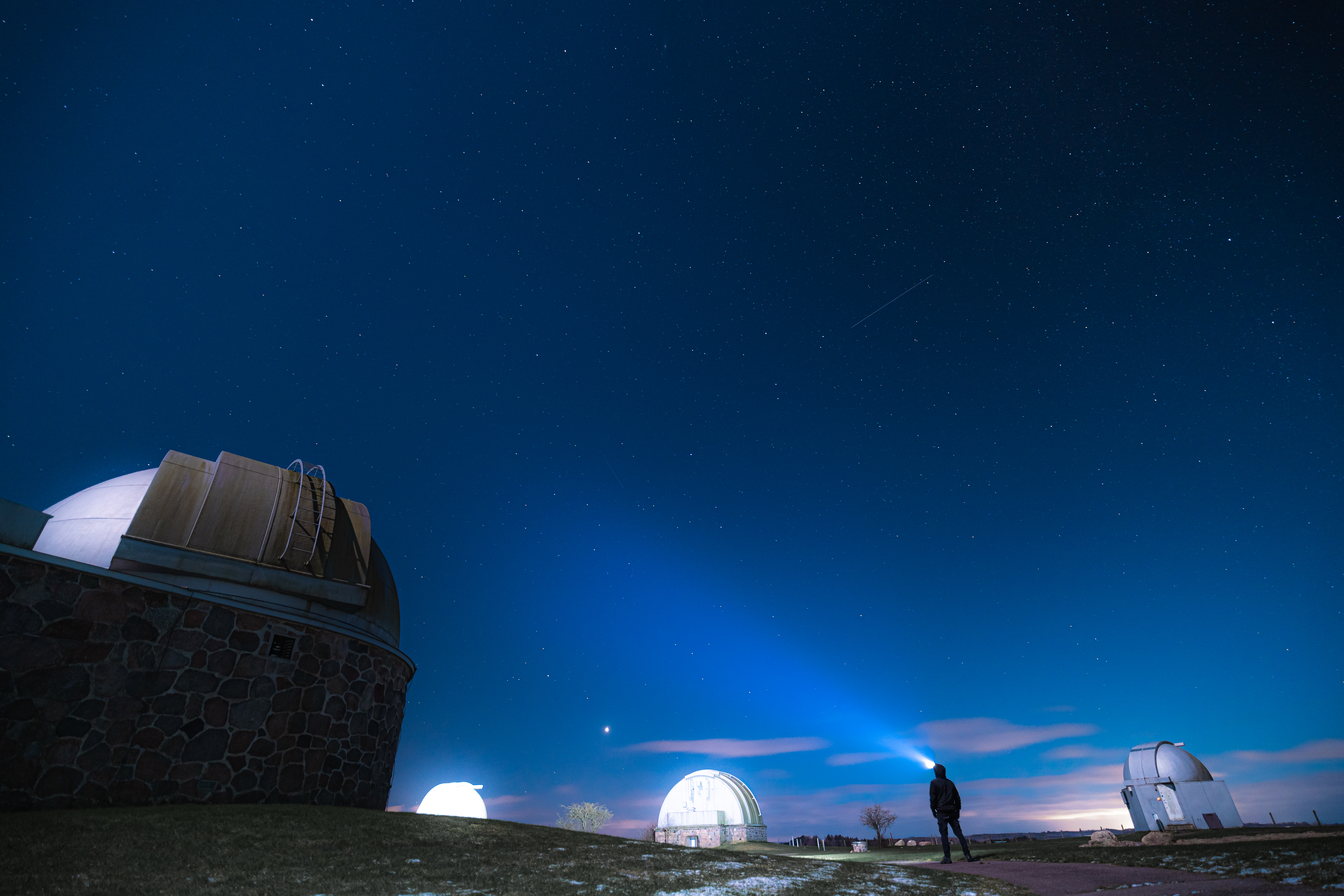 A person standing on a grassy area near observatory domes at night, with a starry sky above and a flashlight beam illuminating the darkness.