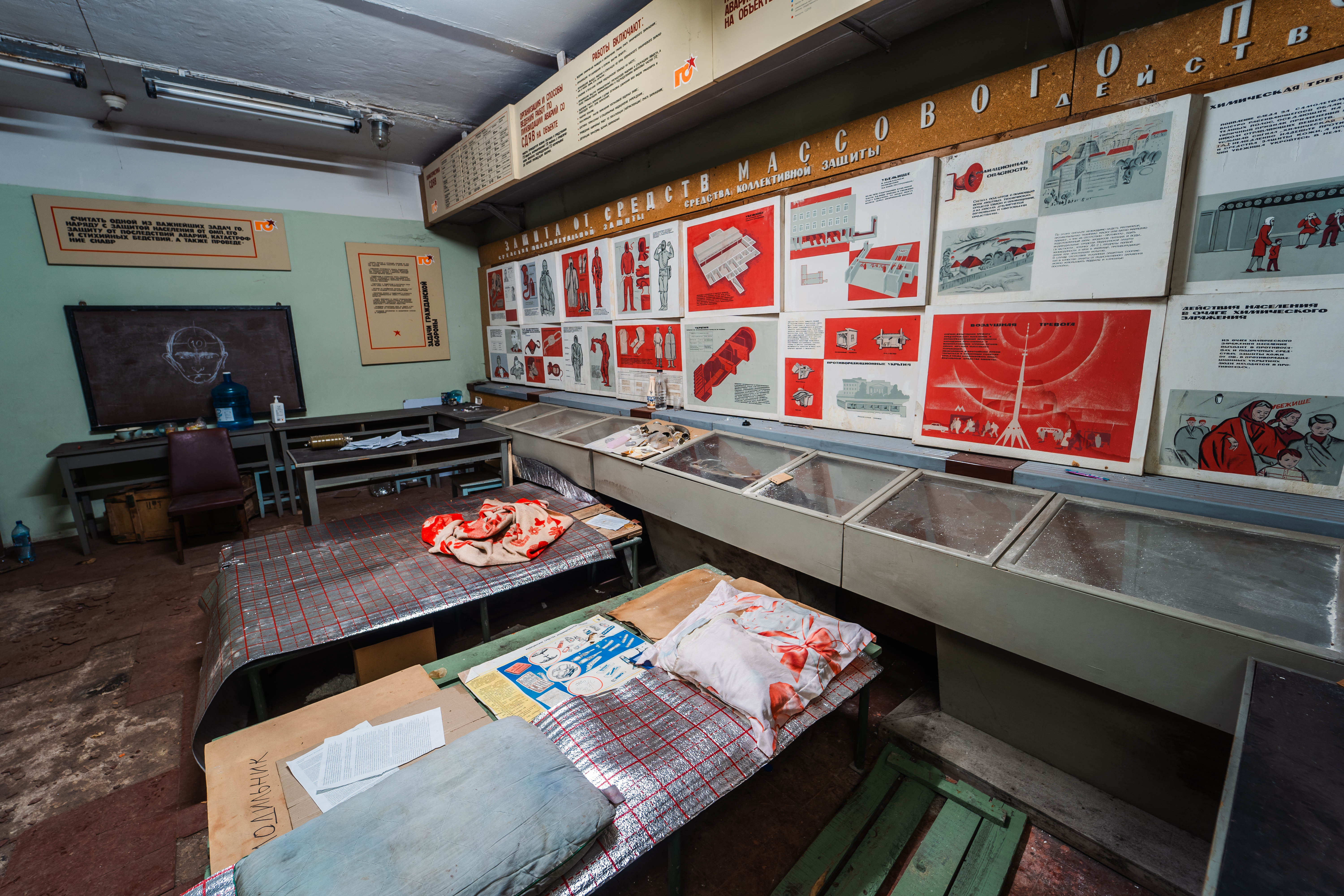 An abandoned classroom with red and white safety posters on the walls, a cluttered table covered with stained fabric, a blackboard, and a water cooler in the corner.