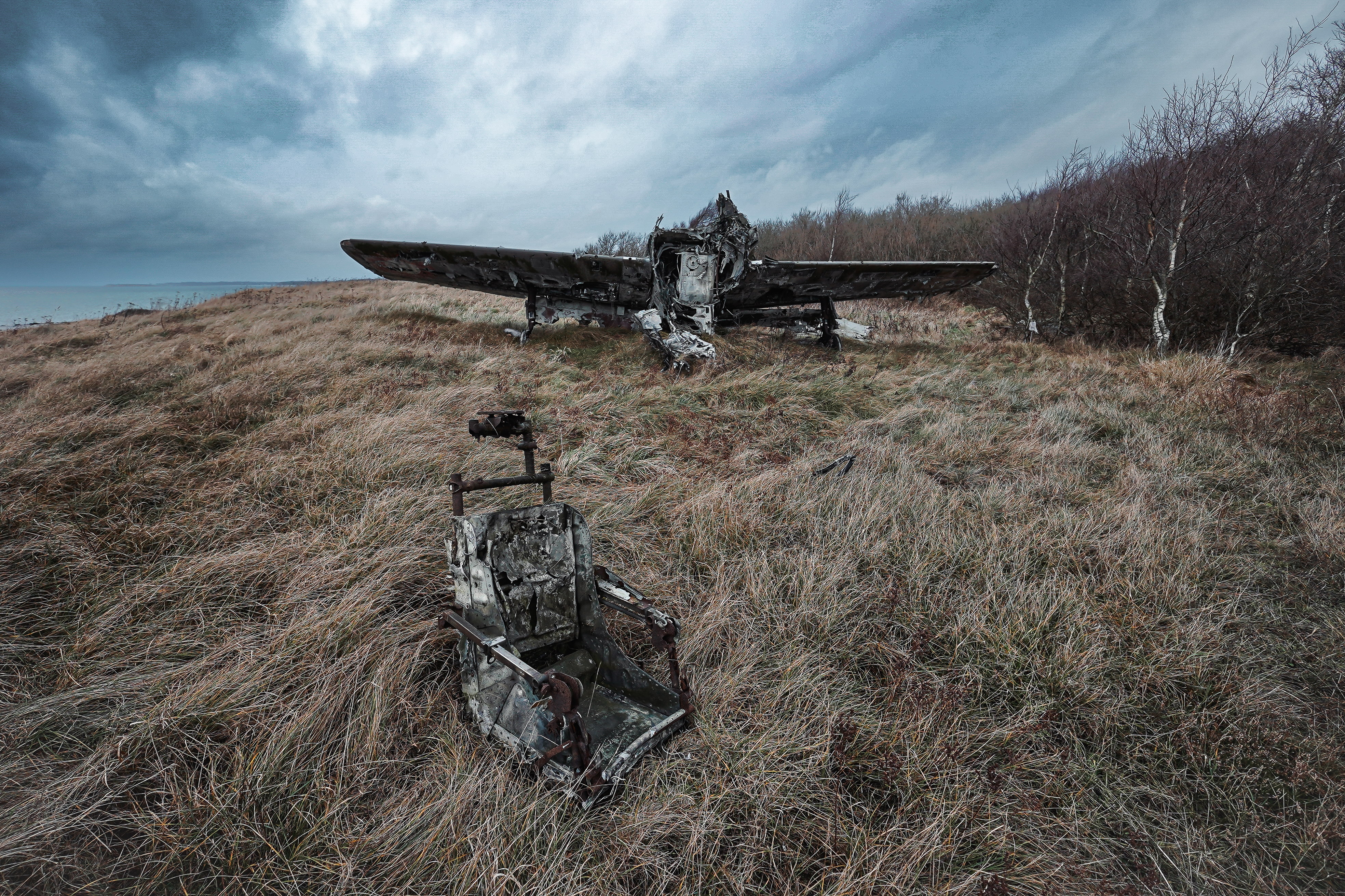 An abandoned aircraft wreck partially visible on a grassy hill, with a rusty pilot's seat in the foreground. The scene is set under a cloudy sky, capturing a desolate and haunting atmosphere.