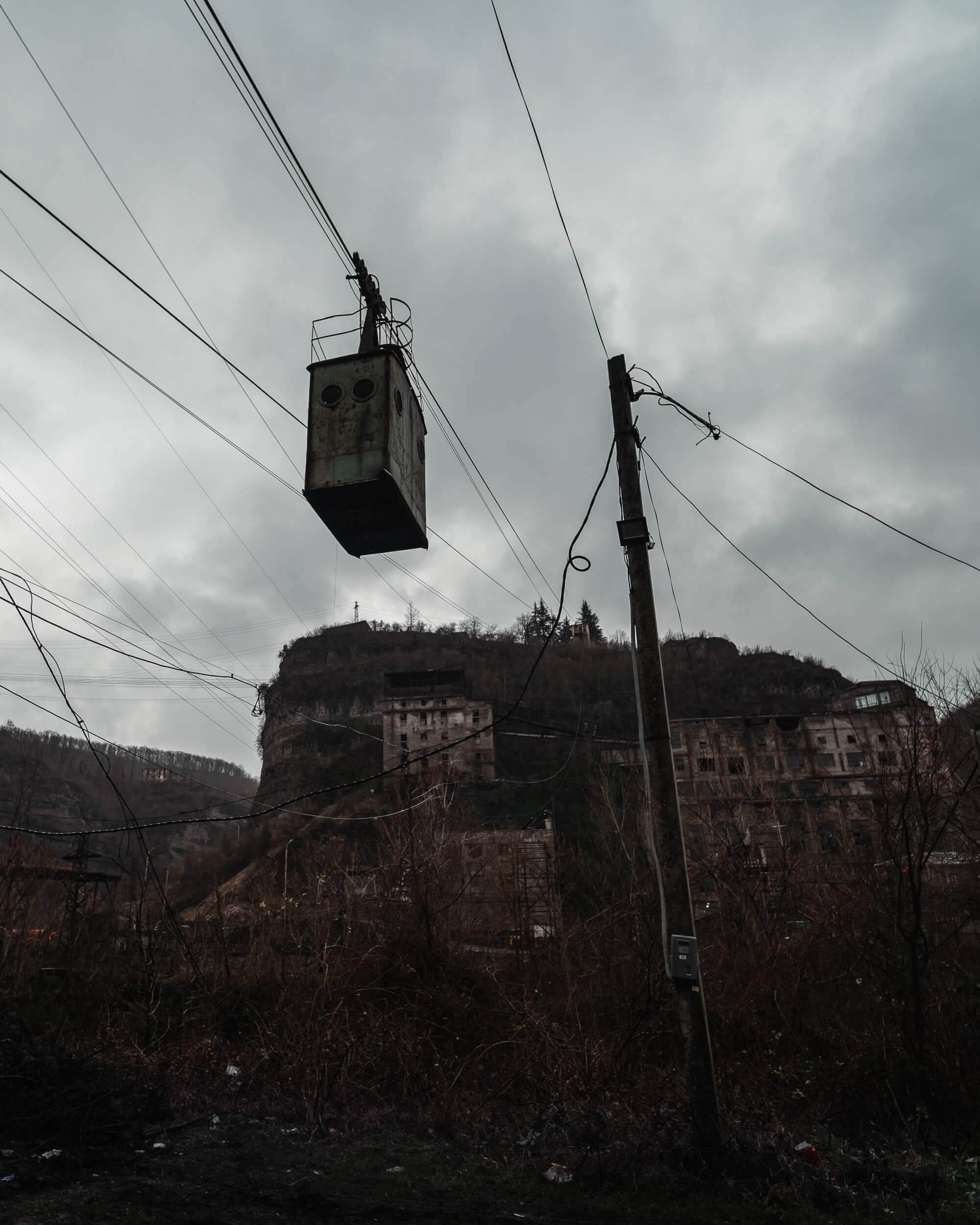 A rusted gondola suspended on power lines with dilapidated buildings on a hillside under a cloudy sky, surrounded by dry vegetation.