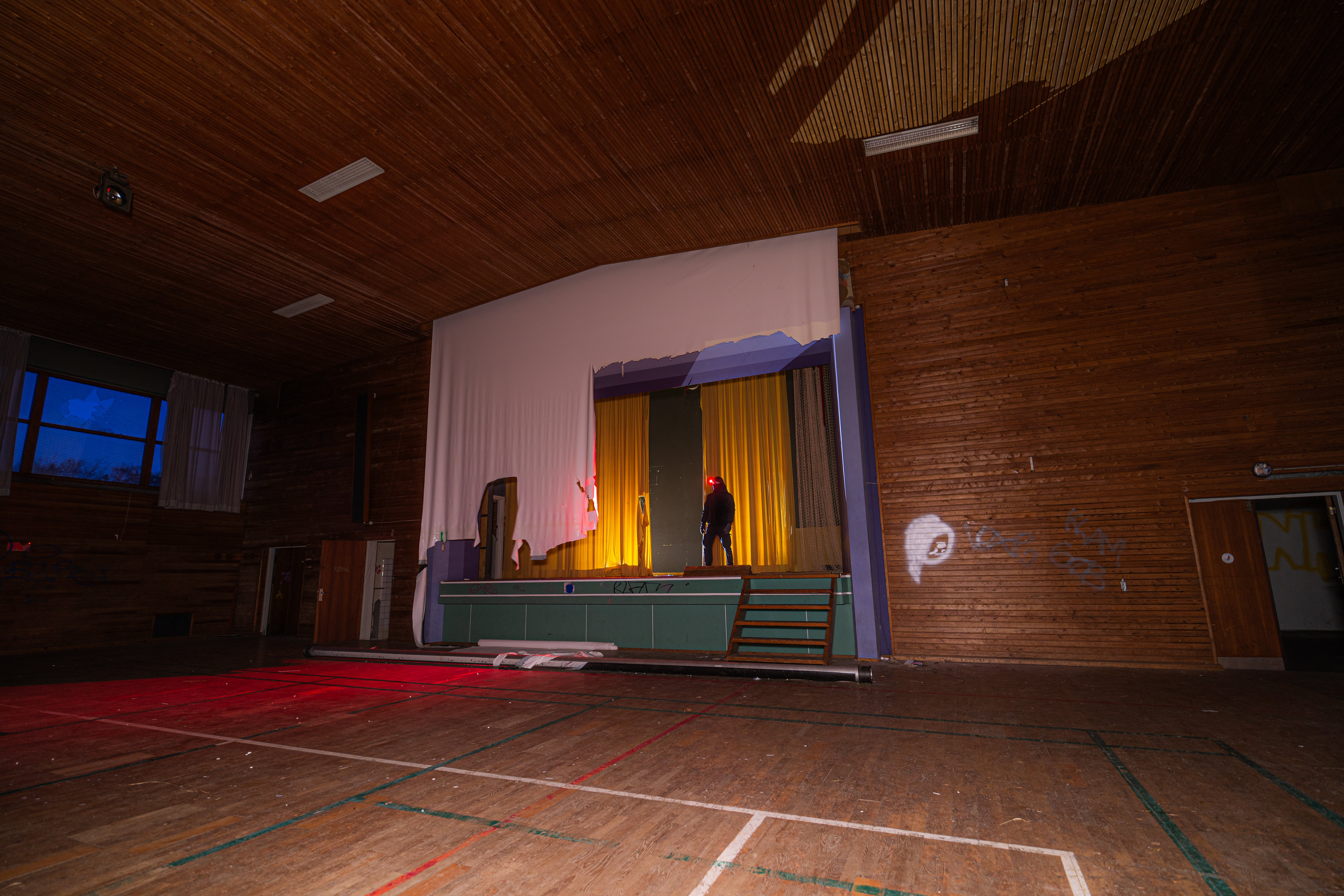 An empty stage in a wooden auditorium. A figure stands under warm yellow lights in front of a draped curtain. Dark wooden walls and a red shadow on the floor create a somber mood.