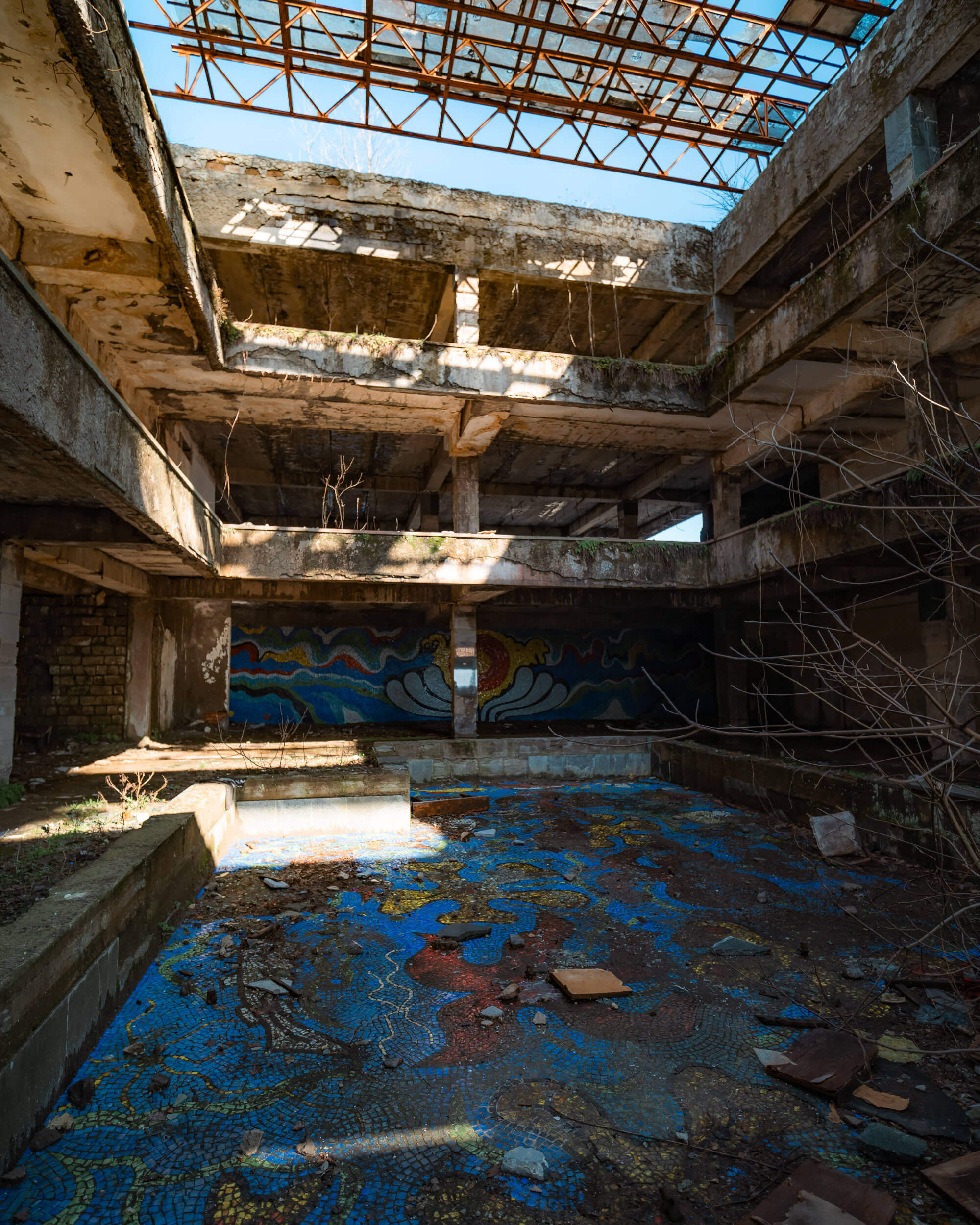 Interior of a ruined building with broken concrete walls, a partially collapsed roof, and colorful mosaic patterns on the floor. Sunlight is streaming in through the roof, illuminating patches of dirt and debris. Sparse vegetation is visible, with vines growing in the cracks.