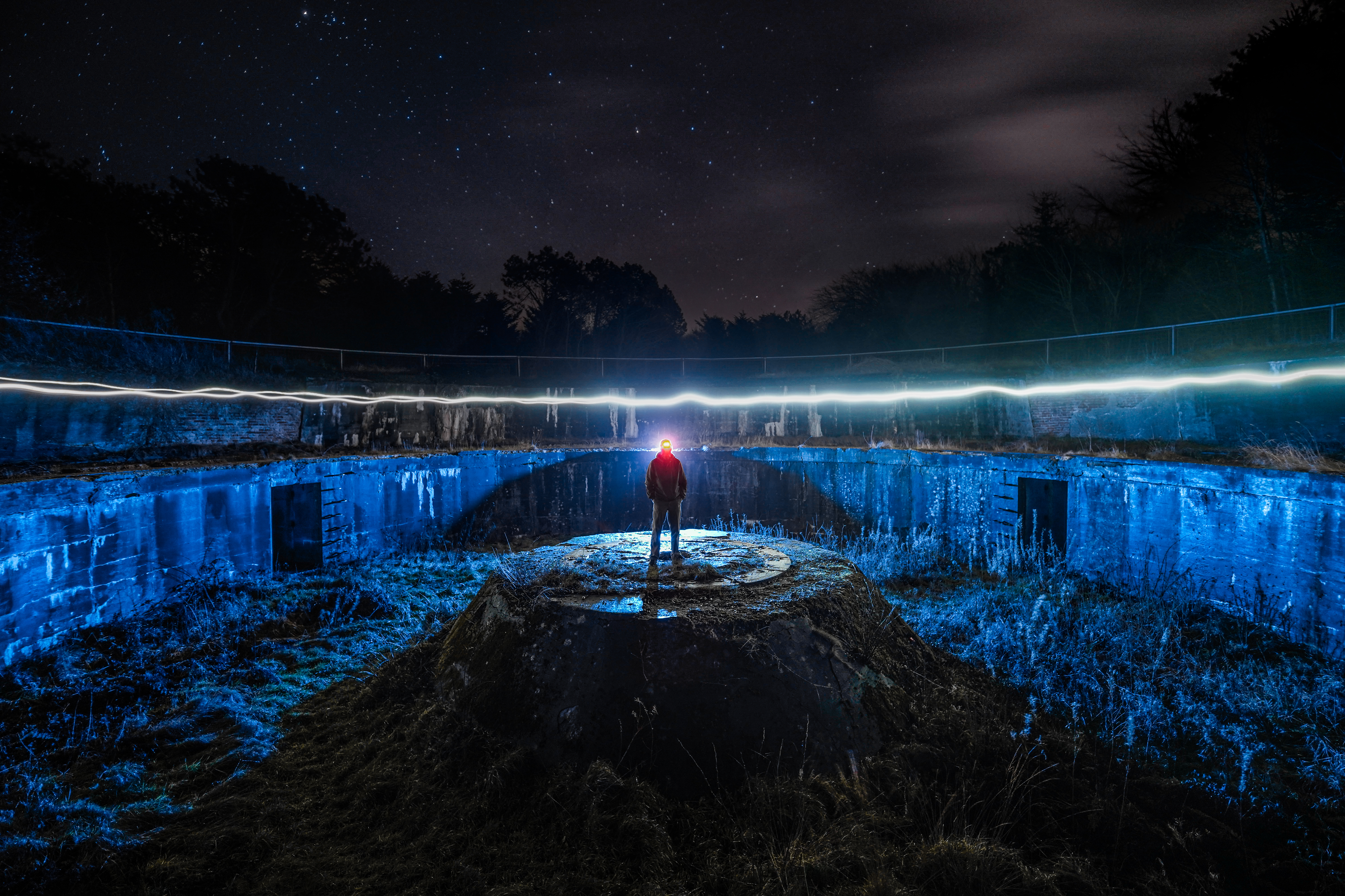 A person stands on an elevated circular concrete structure in a dark landscape, surrounded by blue-lit ground and a starry sky, with a light trail arching above.