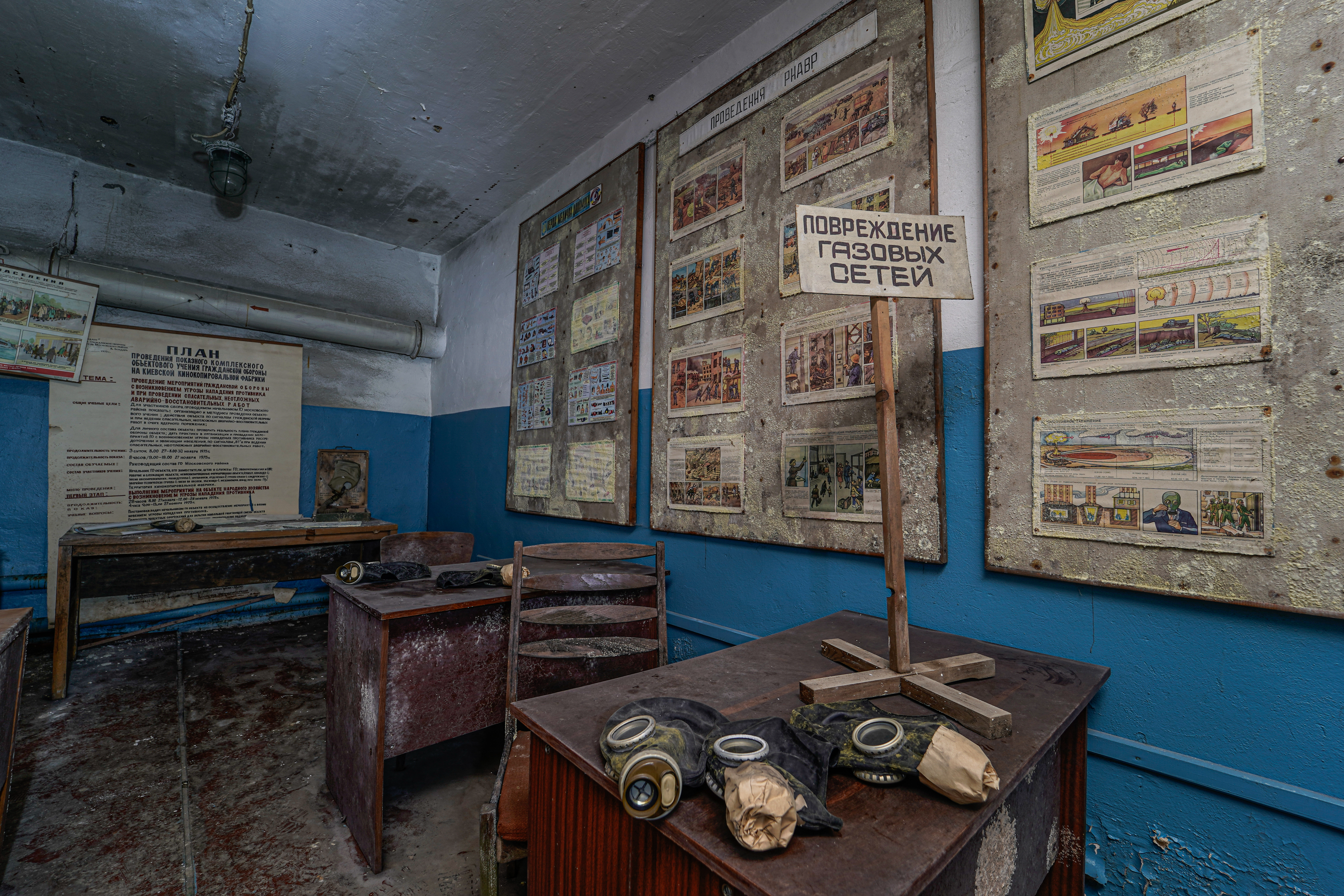 A historical room with damaged walls, various educational posters, a wooden table with old gas masks, and a standing sign in Russian about gas network damage.