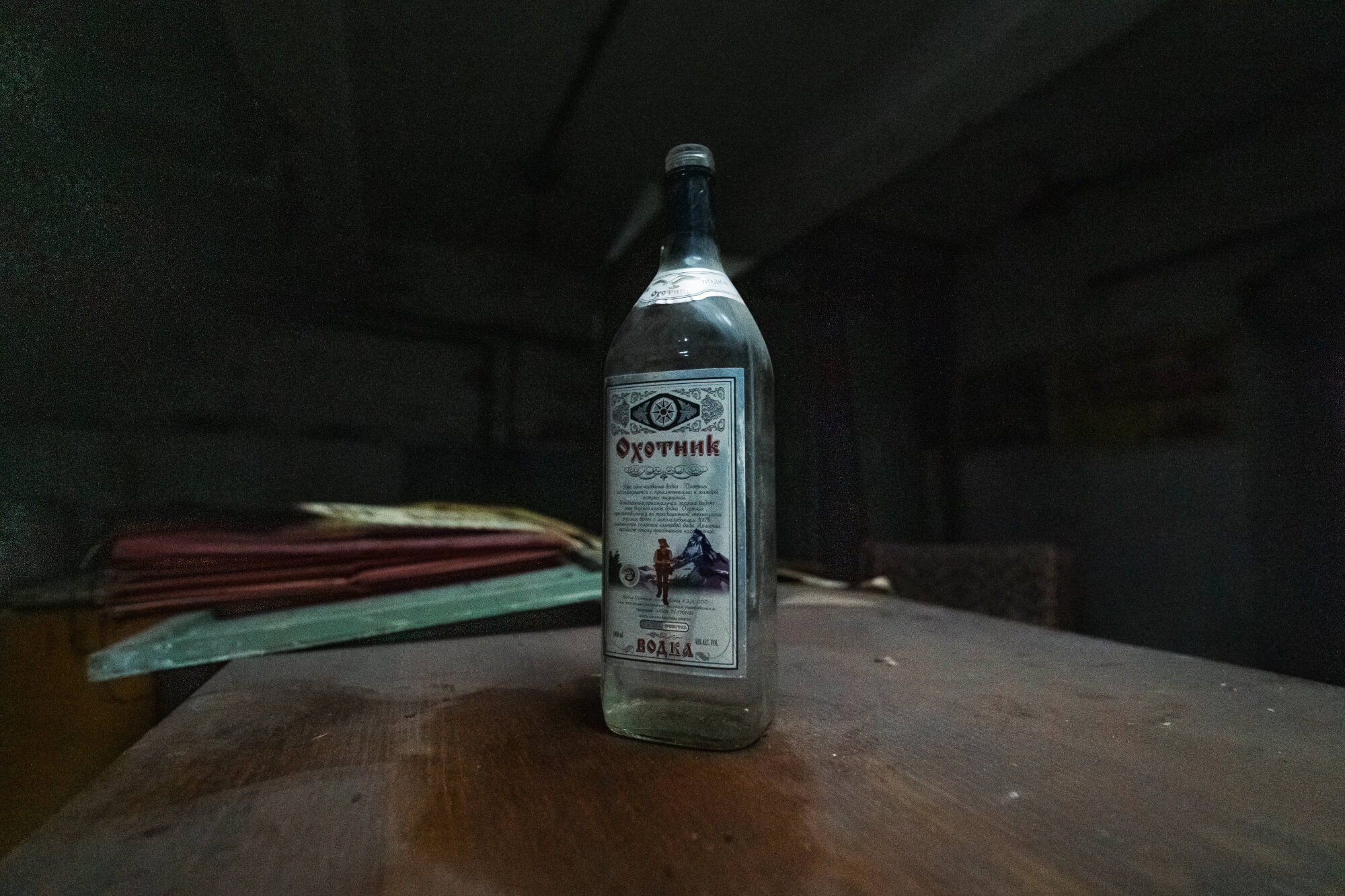 A glass vodka bottle on a wooden table in a dimly lit, abandoned room, surrounded by dusty papers.