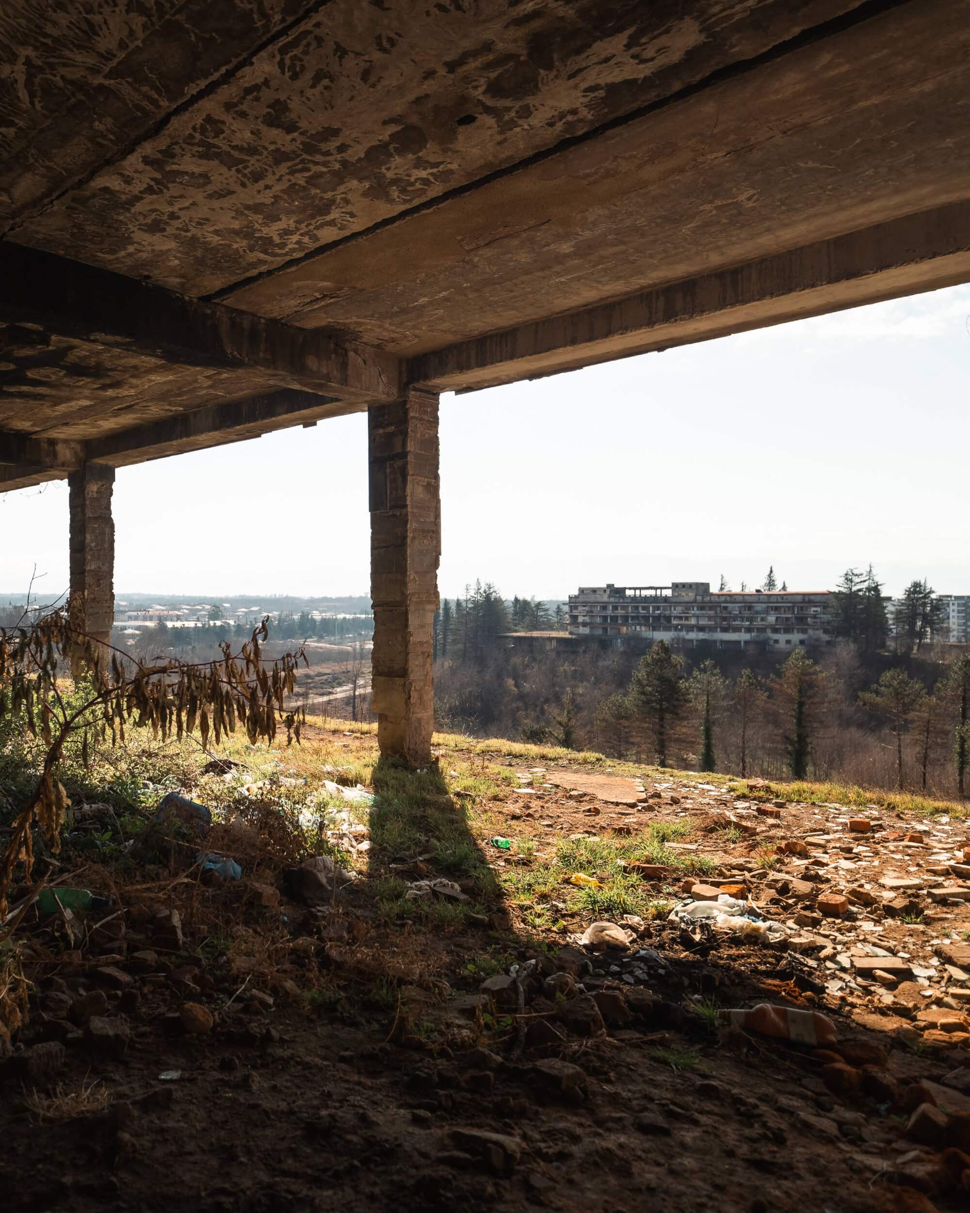 View from an abandoned building, showing concrete pillars and overhanging ceiling above a wild, litter-strewn ground, with an empty, distant structure in the background under a clear sky.