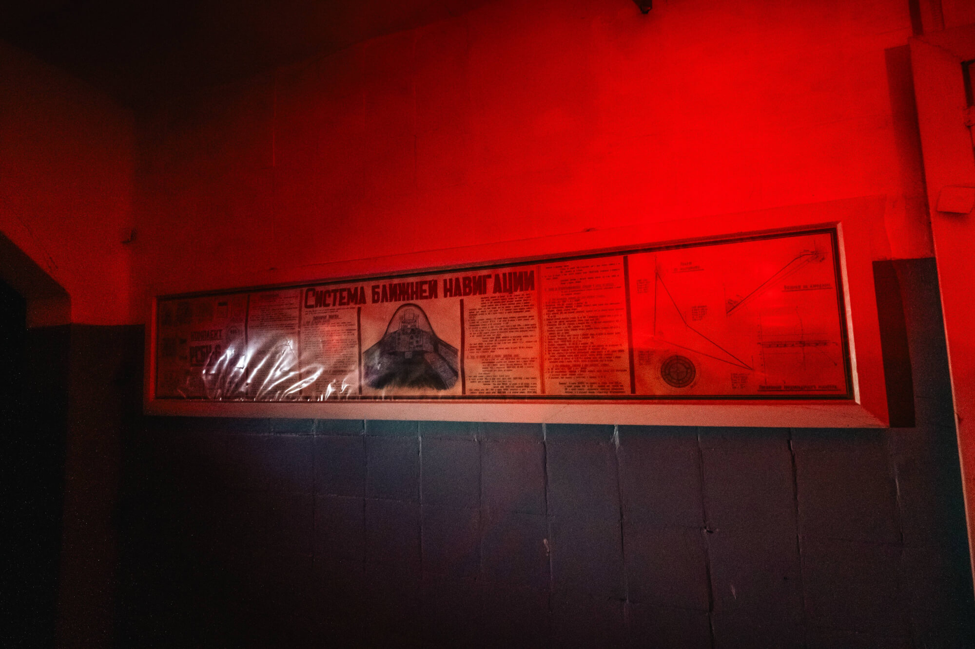 A red-lit wall displaying an informational poster about navigation systems, showing diagrams and text under glass. The surroundings are dim with dark walls, enhancing the dramatic effect.