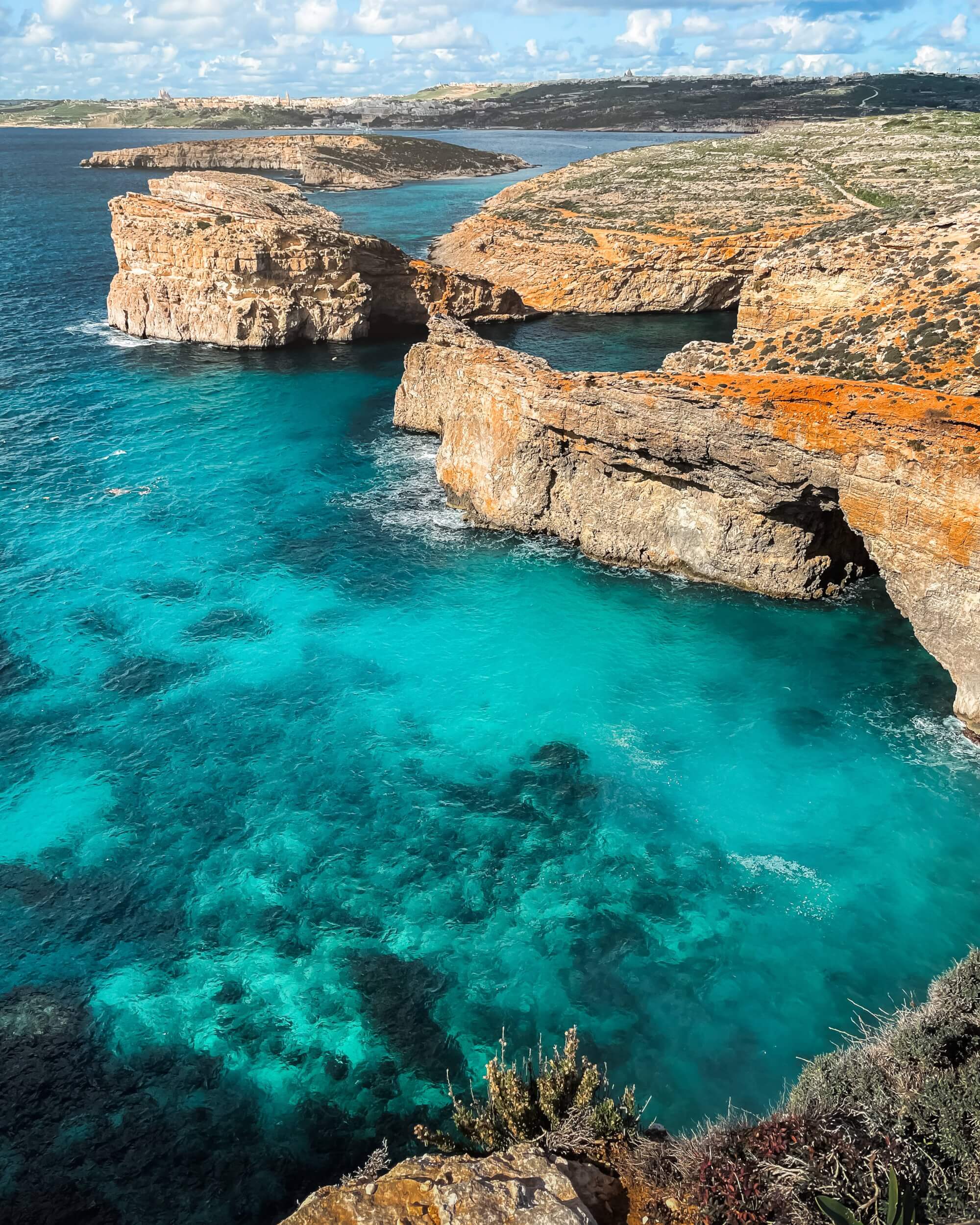 A scenic view of rocky cliffs by a turquoise sea, with clear water showing underwater textures, soft clouds in the sky, and lush greenery along the rocky shore.