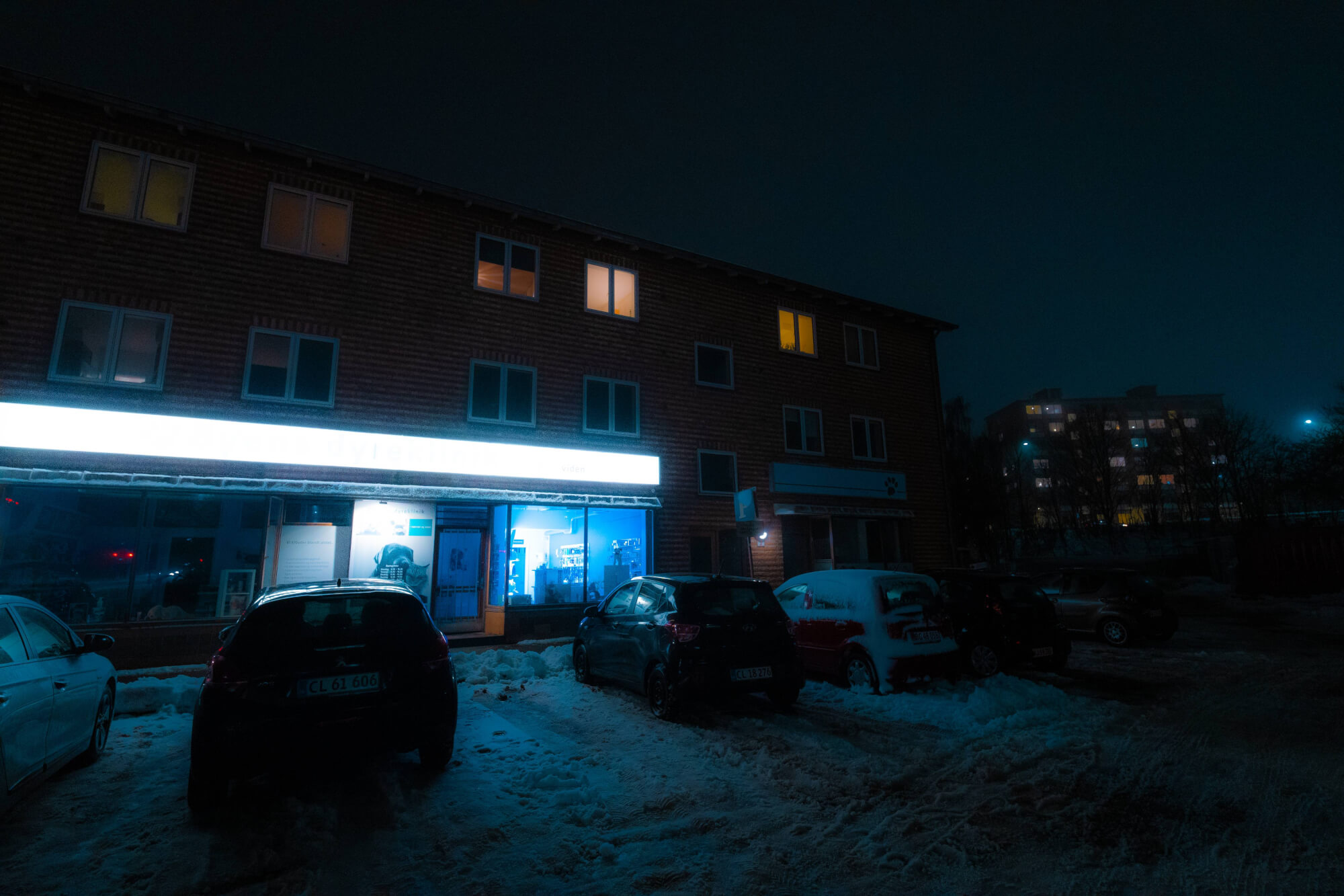 Nighttime scene featuring a snowy parking lot, a partially illuminated brick building, and multiple parked cars. The shop window emits a blue light, and some windows of the building are lit, creating a contrast against the dark sky.