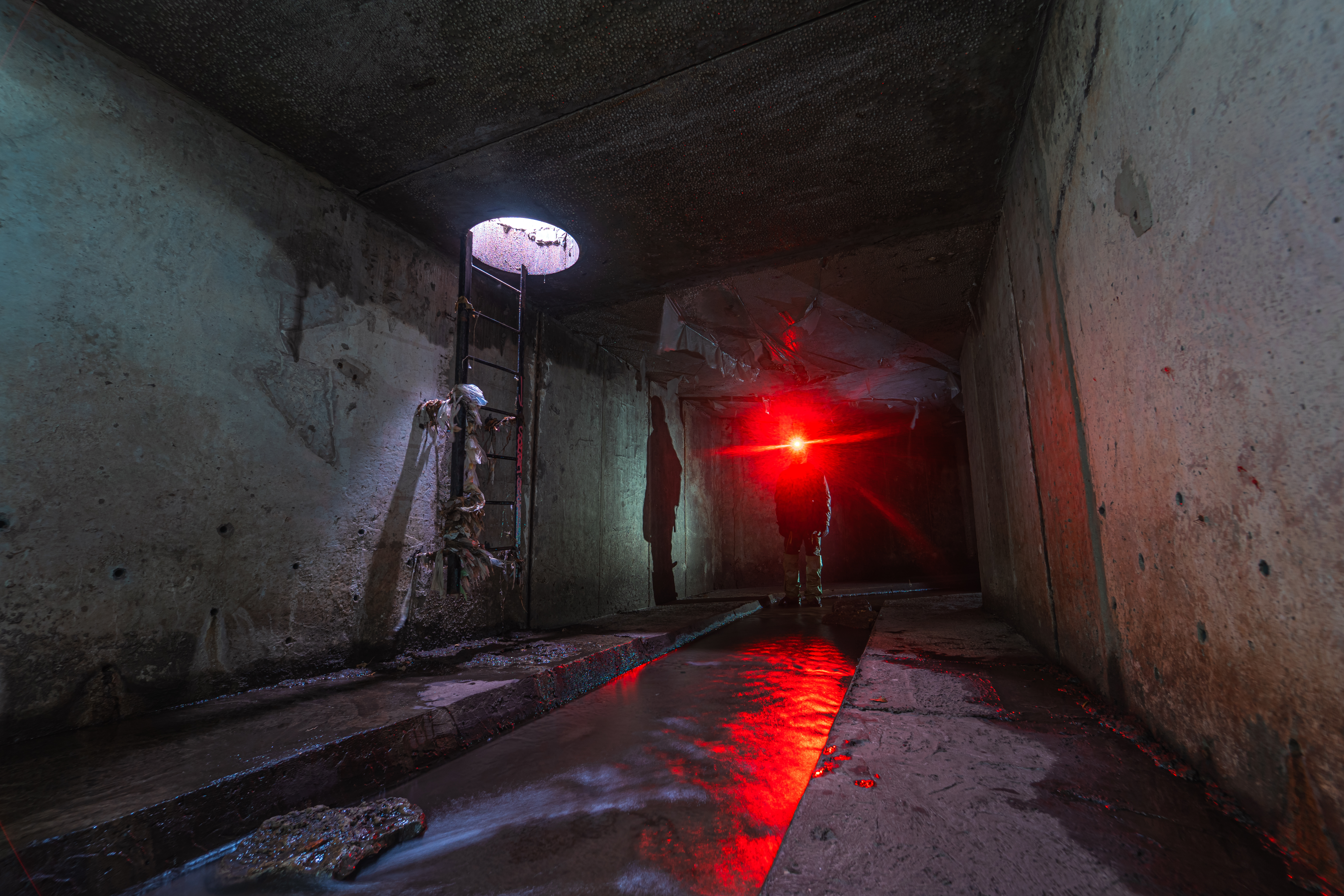 An underground concrete chamber with a ladder on one side leading to a circular opening. A figure stands in the dark, illuminated by a red light, with reflective water on the ground.
