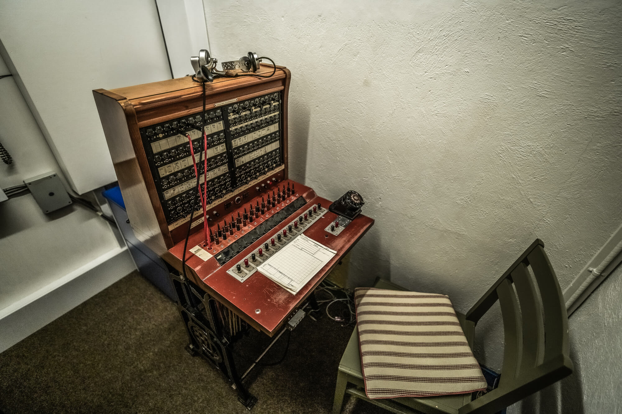 A vintage control panel with numerous buttons, dials, and wires, next to a green chair with a striped cushion, set against a white textured wall.