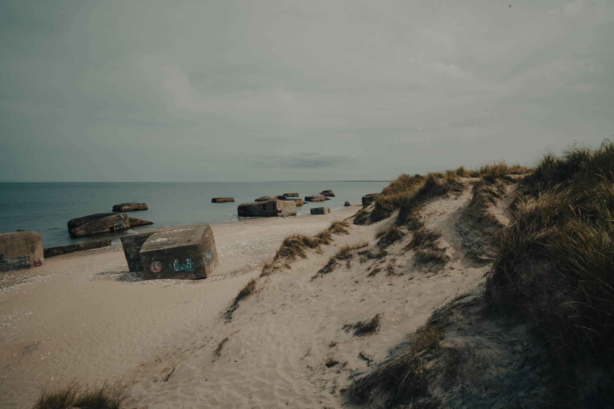 A beach with concrete bunkers scattered along the sand, gentle dunes in the foreground, and a calm sea under a cloudy sky.