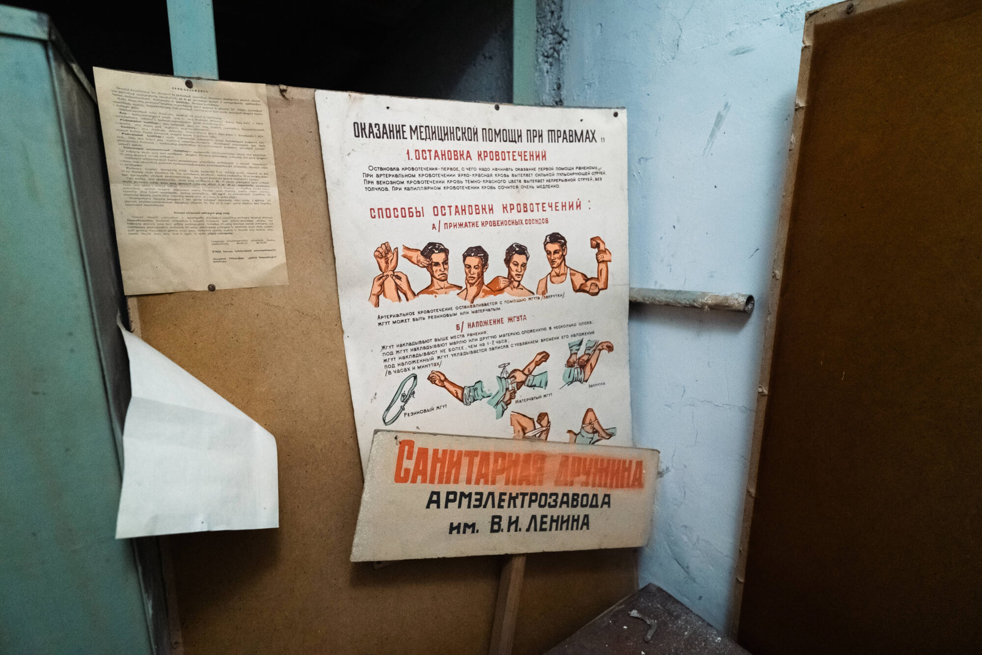 An old medical instruction poster illustrating bleeding control techniques, with accompanying illustrations of muscular arms. A secondary sign in a different style lies below it, against a backdrop of peeling blue walls.
