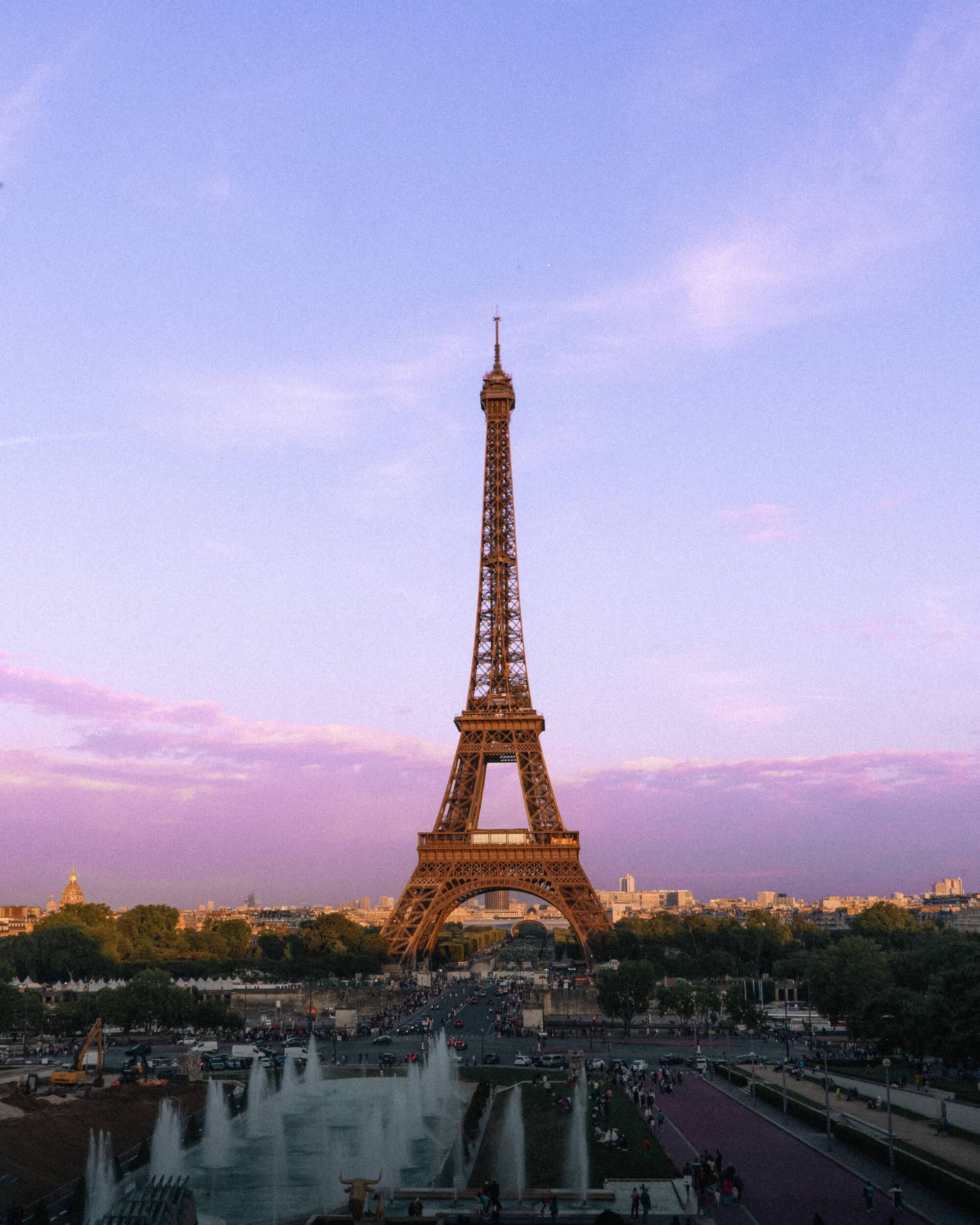 The Eiffel Tower illuminated at dusk with a pastel purple sky, surrounded by trees and a lively crowd near fountains.