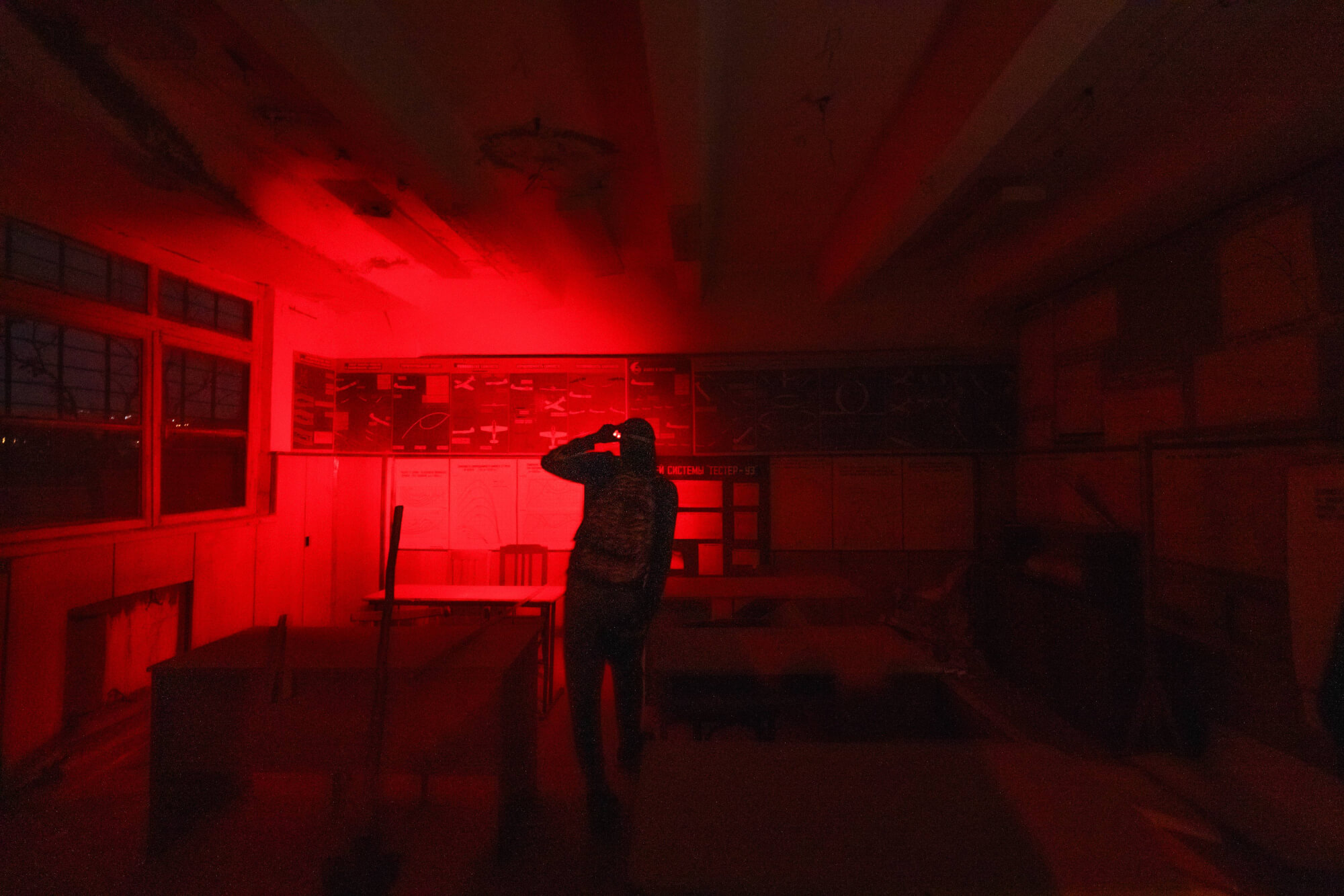 A dimly lit abandoned classroom illuminated by red light. A silhouetted figure stands in the middle, turned away, with faded educational charts on the wall and broken windows in the background.