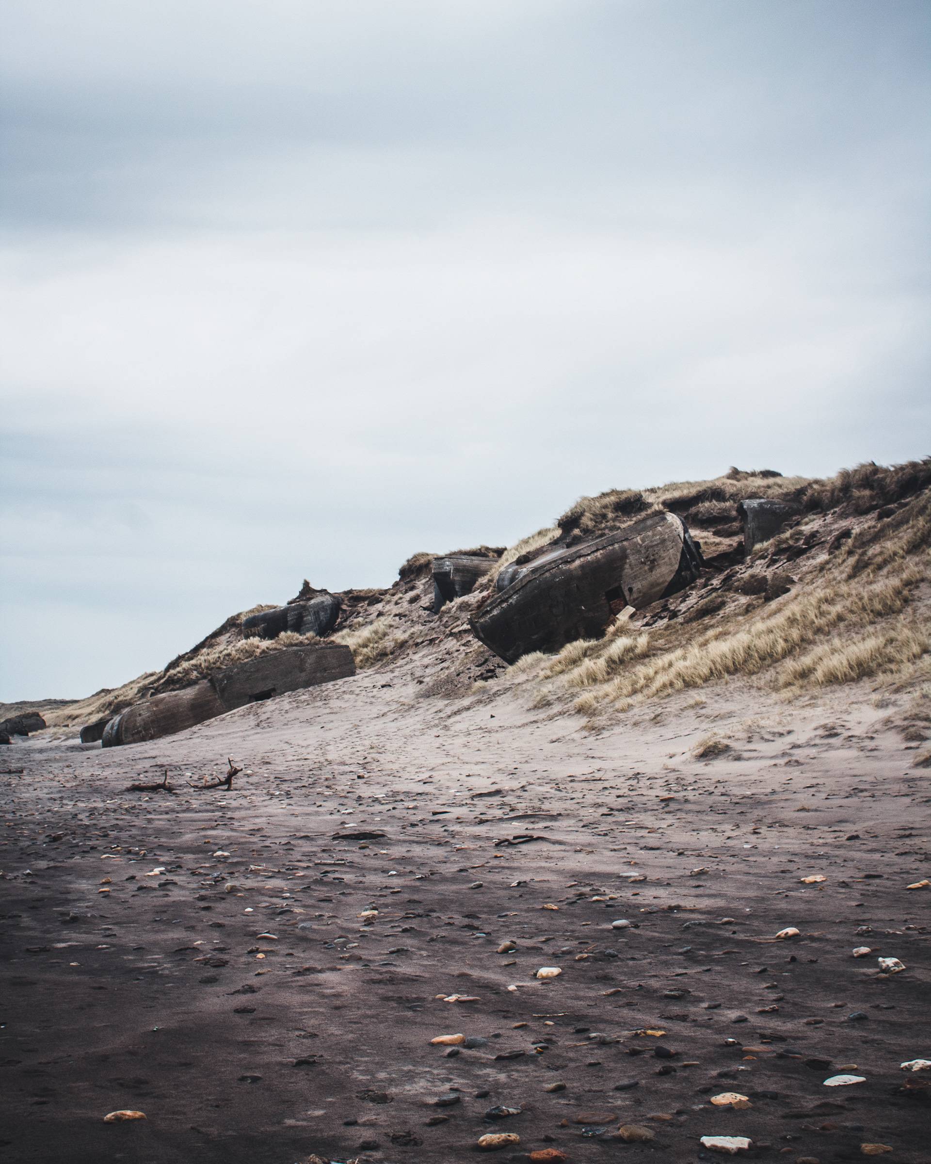 A desolate beach scene featuring a sandy shore with scattered stones, old structures on a hillside, and a cloudy gray sky.