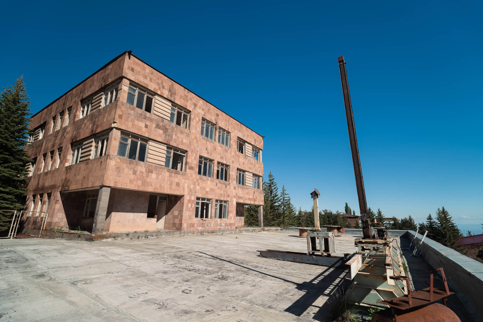 A large, abandoned building made of rough stone with broken windows, set against a bright blue sky. In the foreground, there are remnants of machinery and scattered metal parts, with trees visible in the background.