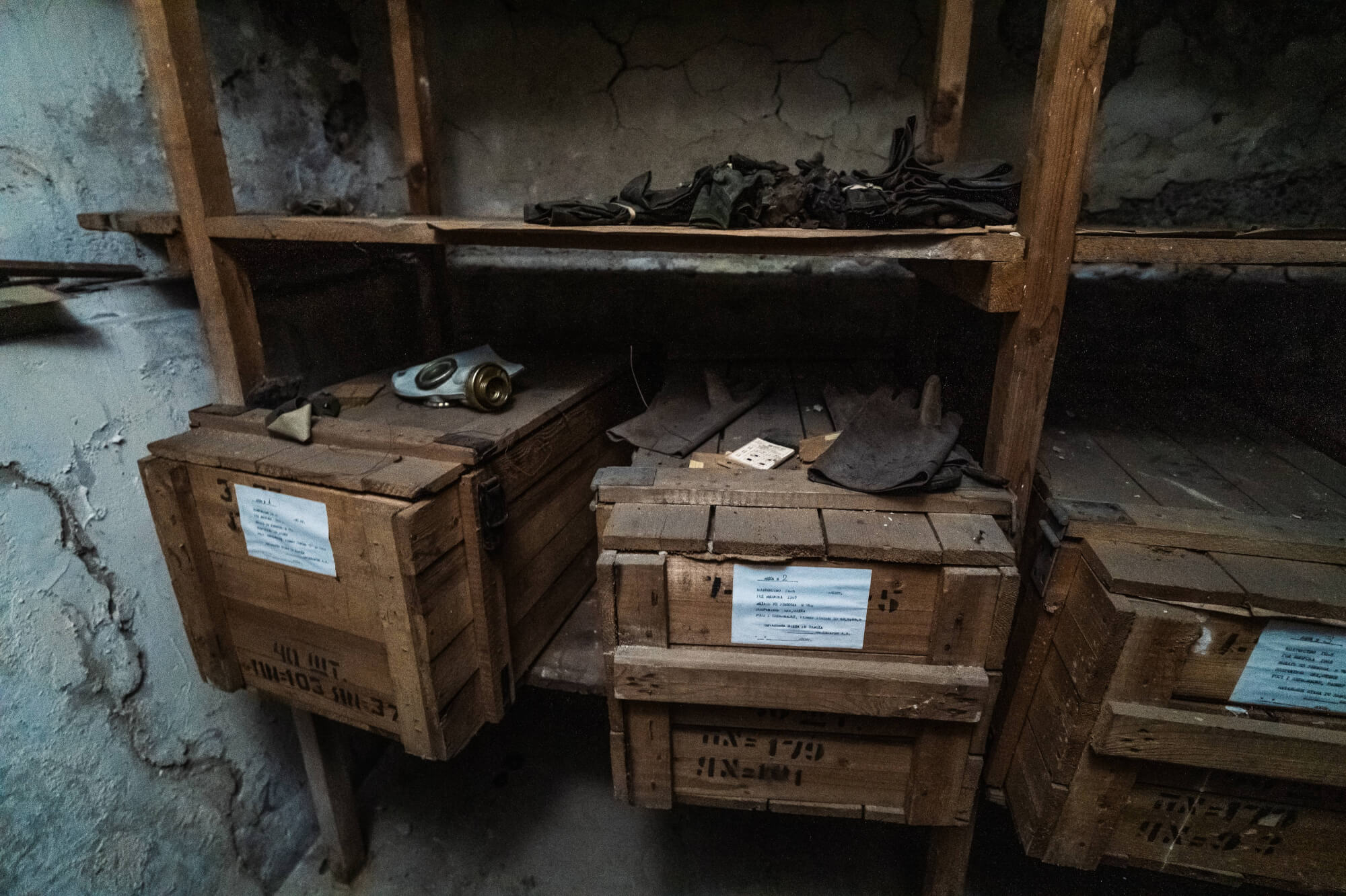 A shadowy storage room with wooden shelves containing wooden boxes, work gloves, and a gas mask. The background shows cracked walls, enhancing the atmosphere of neglect.