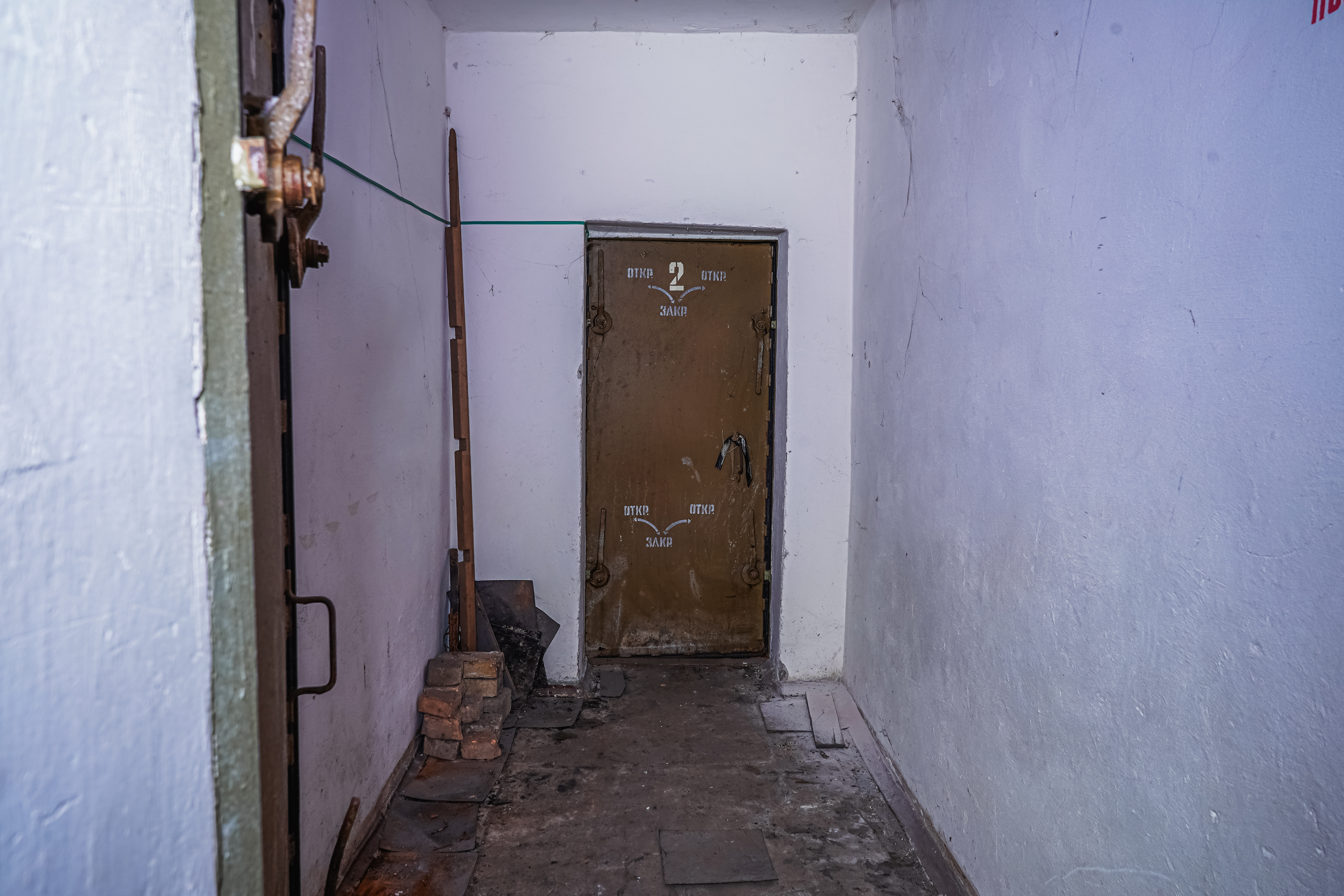 A narrow corridor with a weathered brown door labeled '2' at the end. The walls are white and slightly worn, with a wooden stick leaning against the left wall. There are uneven tiles on the floor and a pile of bricks in the corner, creating an abandoned and mysterious ambiance.