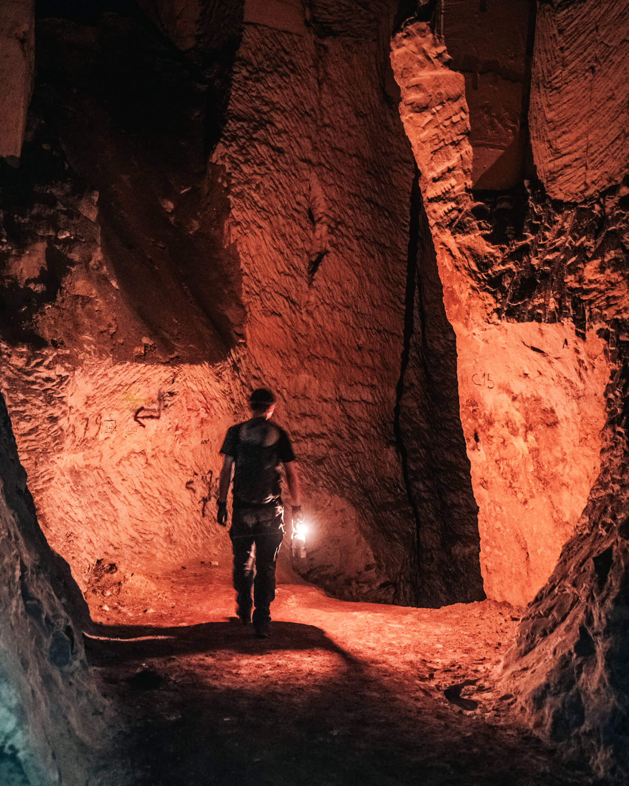 A person walking through a dimly lit cave, holding a lantern that casts a warm glow on the rocky surroundings. The cave walls are irregular and textured, with shades of red and orange illuminating the scene.