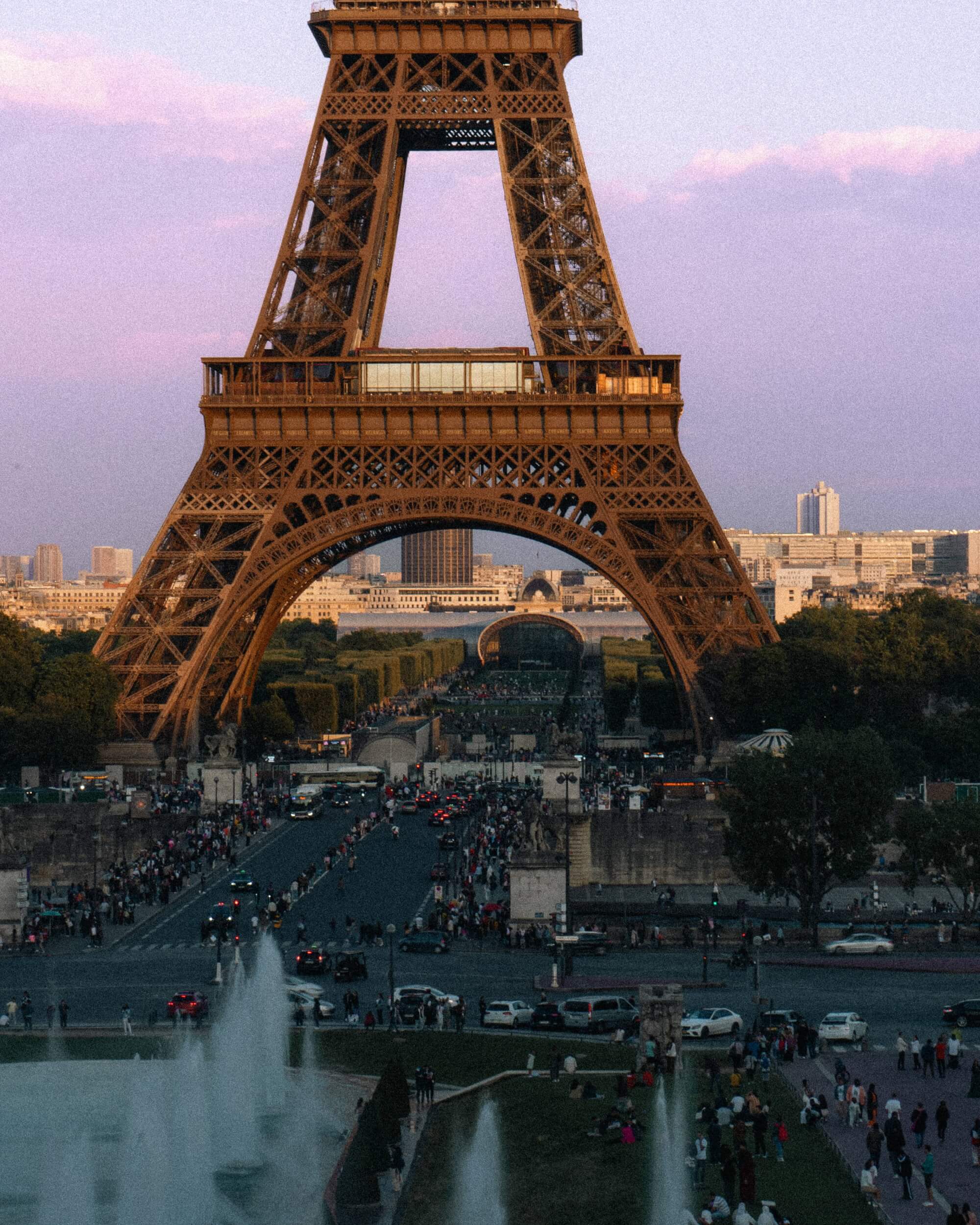 A view of the Eiffel Tower during twilight, with a crowd of people along a pathway and traffic moving on the street below. The sky features pastel colors as the day transitions to night.