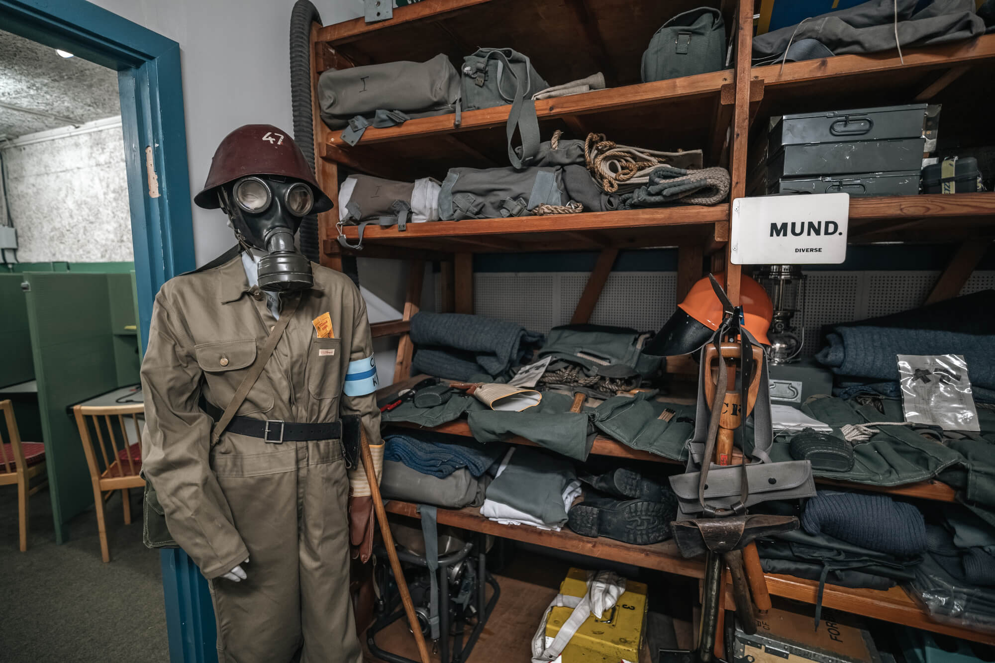 A mannequin in a gas mask and military uniform stands in a room filled with shelves lined with military gear and supplies, including bags, uniforms, and tools. The setting is dimly lit, creating a somber mood.