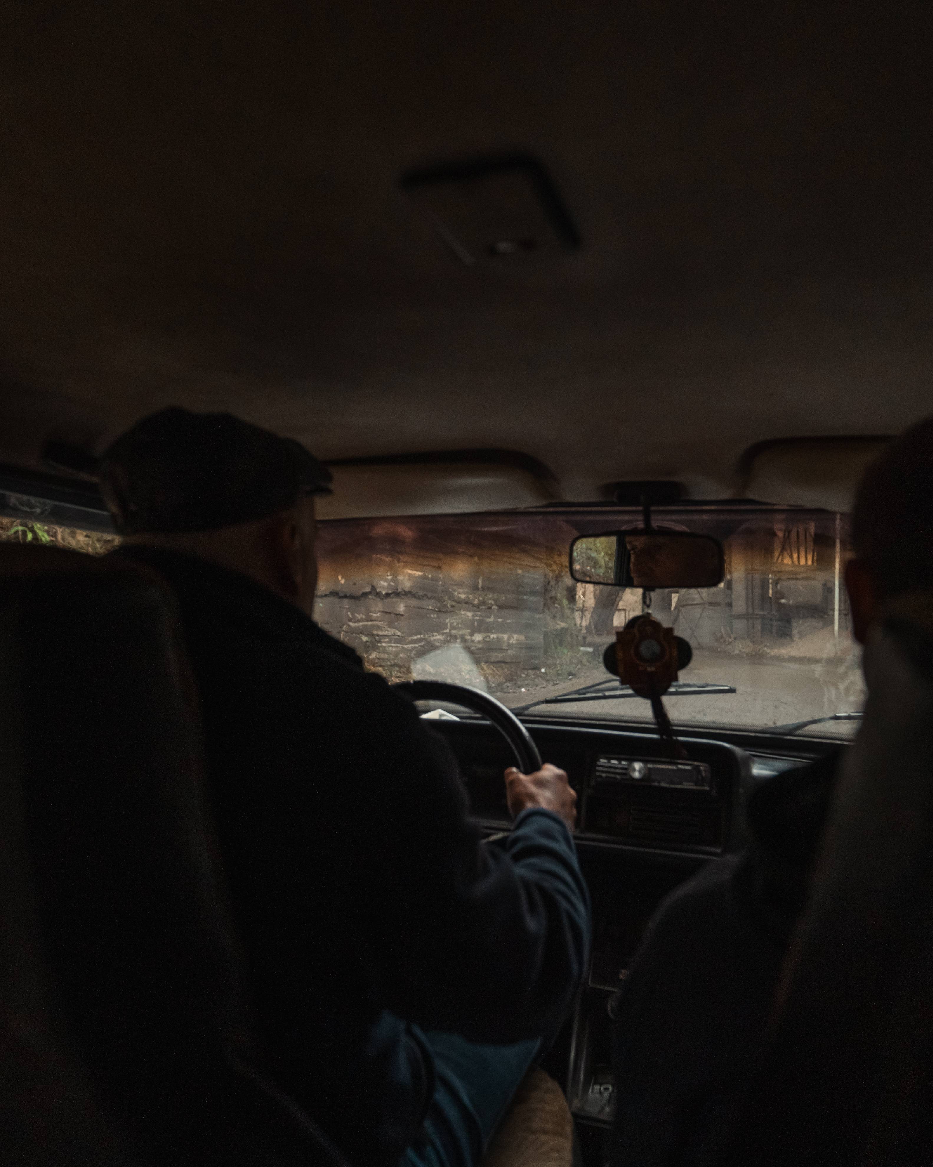 View from inside an old car, showing a driver and a passenger. The driver, wearing a cap, is focused on the road, while the passenger looks in the rearview mirror. The scene outside features blurred trees and a gravel pathway.