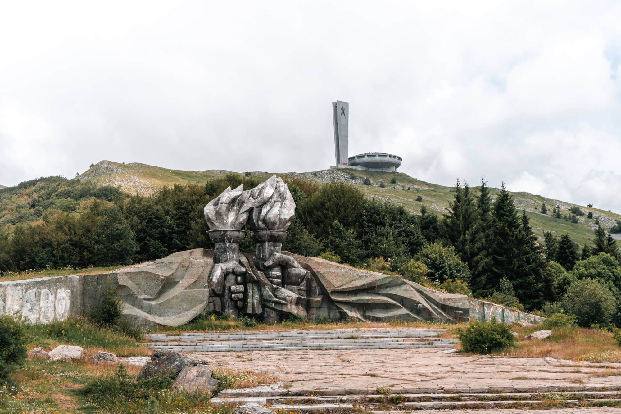 A large sculpture of hands holding flames in the foreground with a modern, circular building and a tall monument in the background, set on a grassy hillside under a cloudy sky.