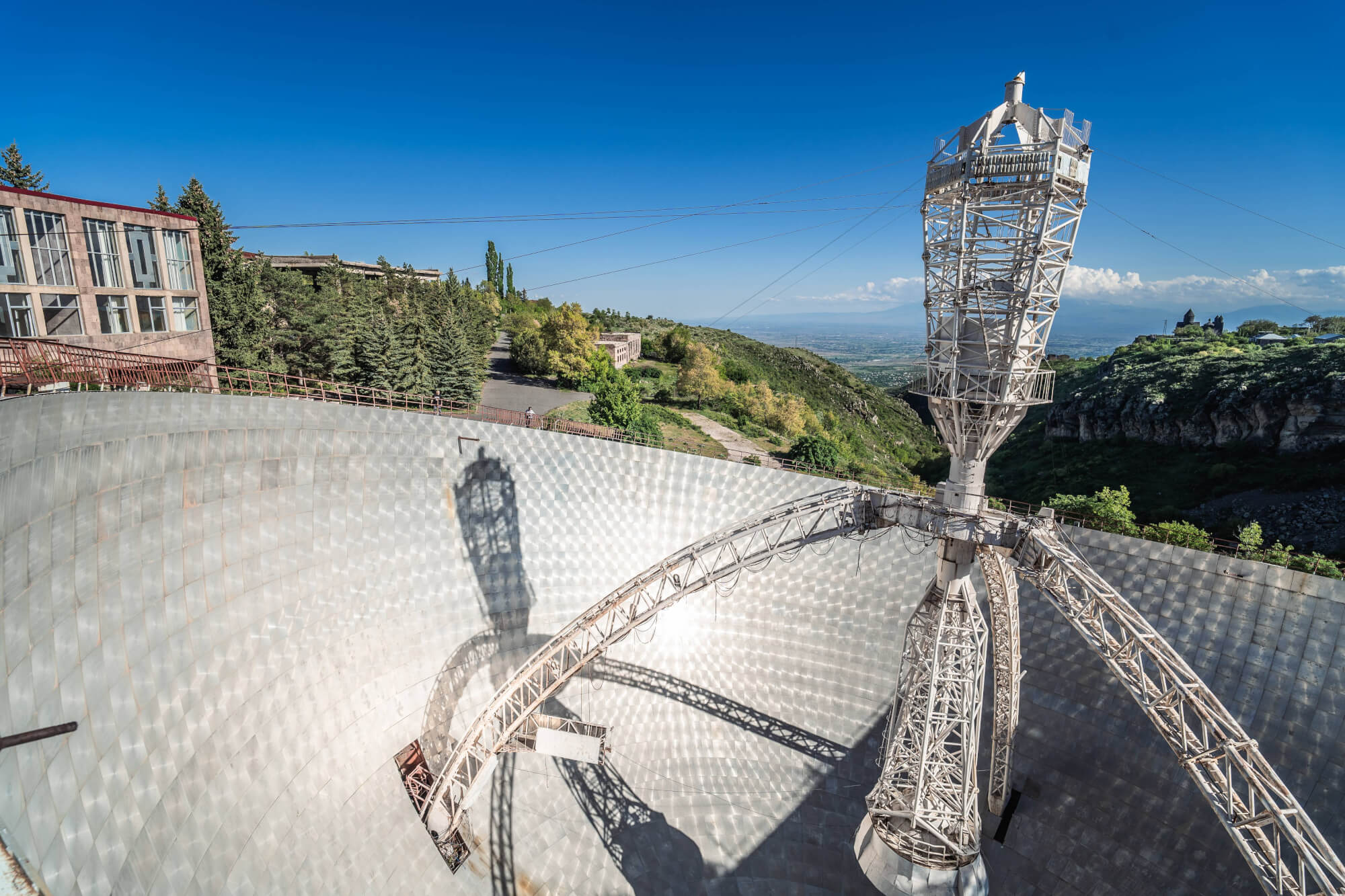 A large metallic dish structure with a curved surface and intricate framework. In the background, green hills and a clear blue sky create a serene landscape. The shadows of the structure are visible on the dish.