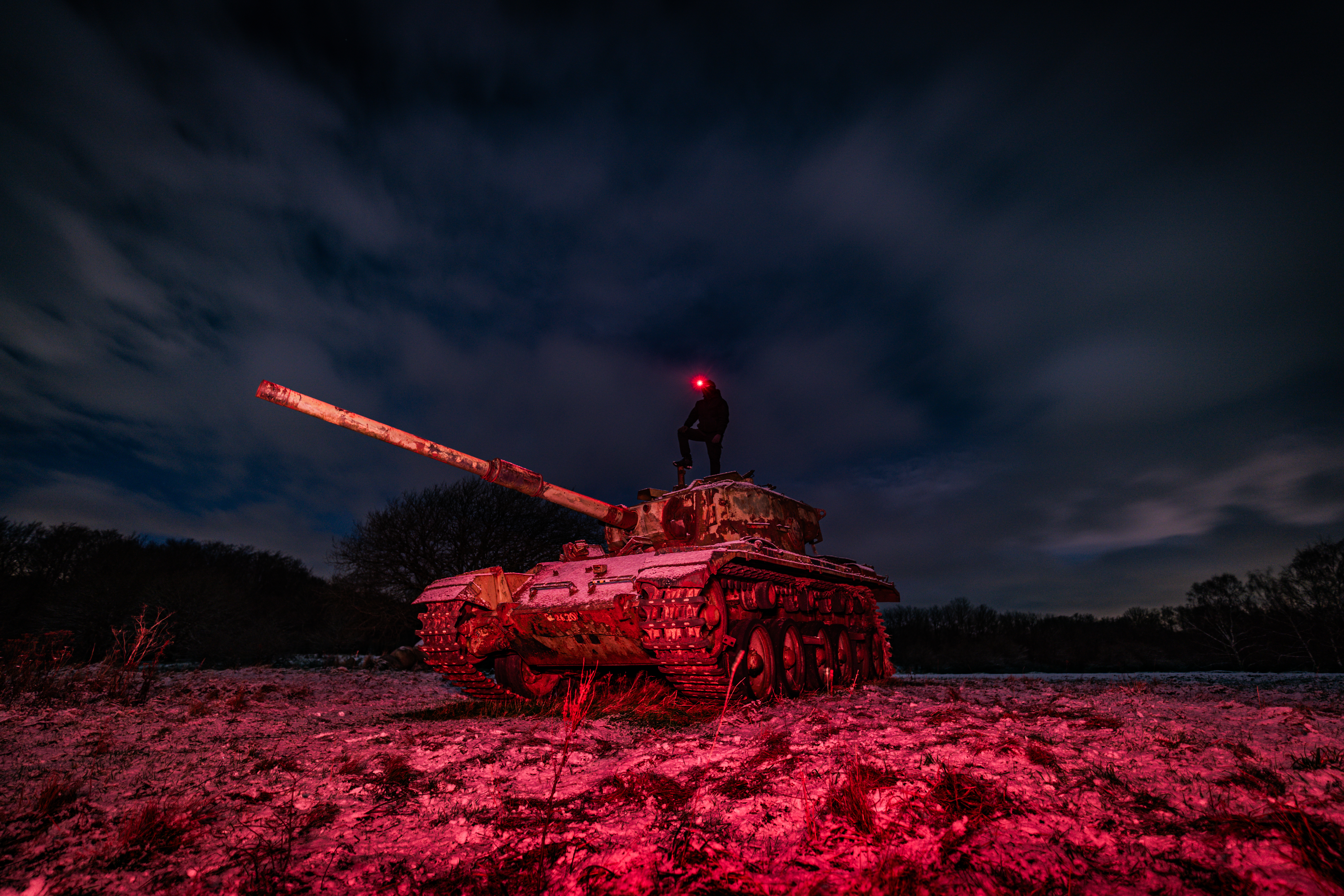 A rusted military tank on a snowy ground at night, with a person standing on top of it, wearing a red light on their head. The sky is dark with scattered clouds, and the tank is illuminated in red light.
