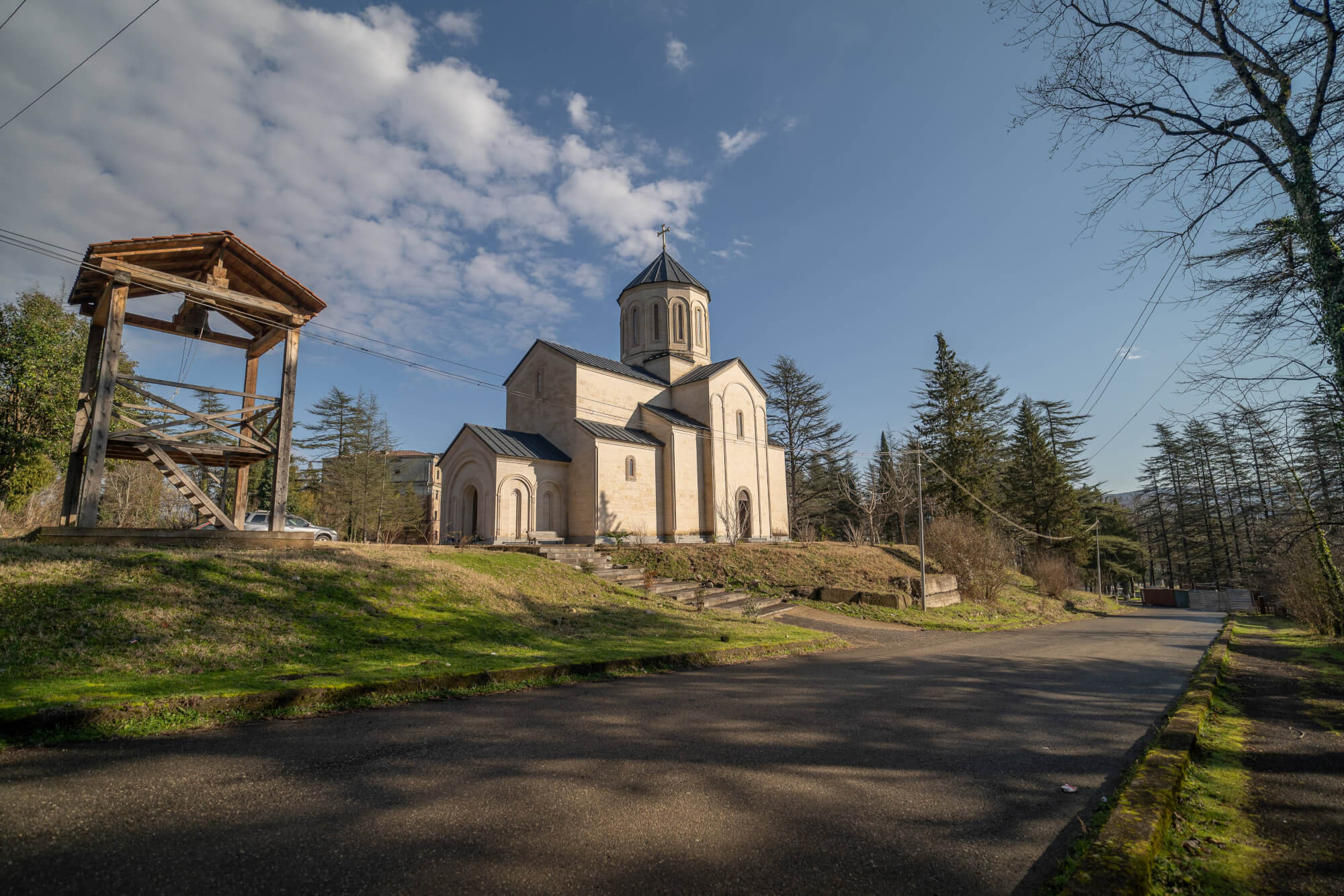 A beautiful church with a dome and arches, set against a blue sky and clouds, with a wooden bell tower to the left and trees in the background. A road leads up to the church.